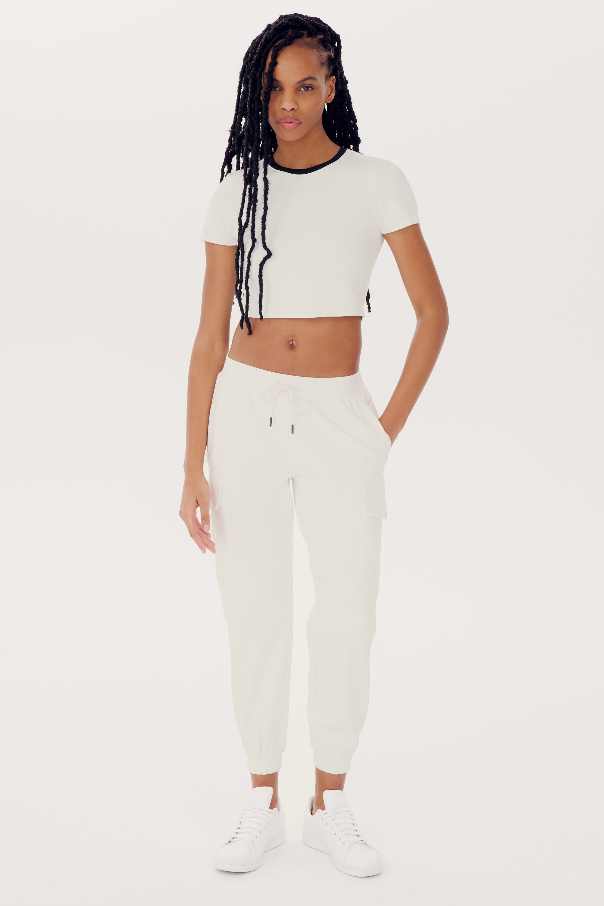 A person with long braided hair, wearing a SPLITS59 Airweight S/S Crop - White/Black and white jogger pants made of spandex, along with matching white sneakers, stands facing the camera with one hand in their pocket.