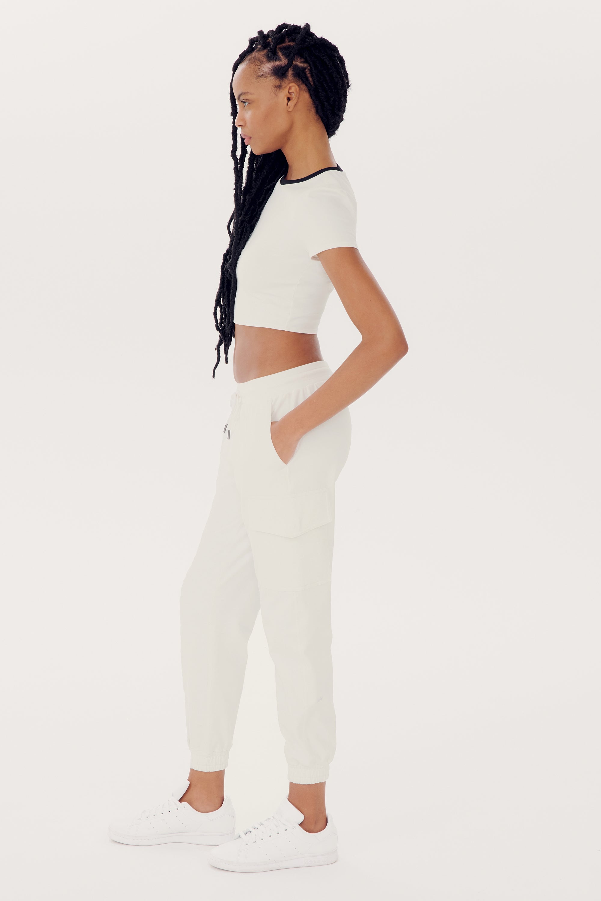 A woman with long braided hair wears an Airweight S/S Crop - White/Black by SPLITS59, white jogger pants made of spandex, and white sneakers, standing in profile against a plain white background.