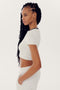 A person with long braided hair is standing in profile, wearing a short-sleeved white Airweight S/S Crop - White/Black by SPLITS59 and matching nylon pants, with one hand in their pocket.