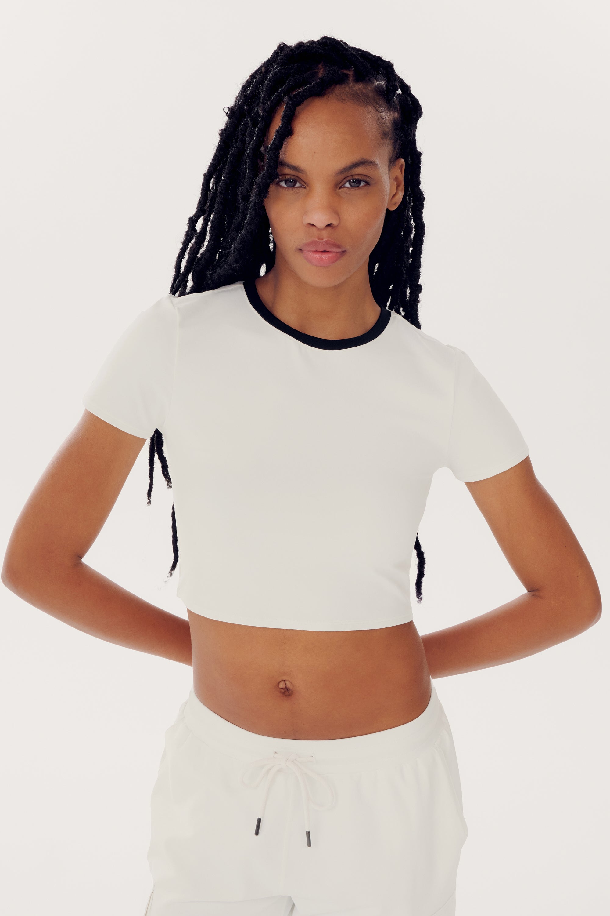 Person with long braided hair wearing a SPLITS59 Airweight S/S Crop - White/Black and white pants made from a mix of nylon and spandex, standing with hands on hips against a plain background.