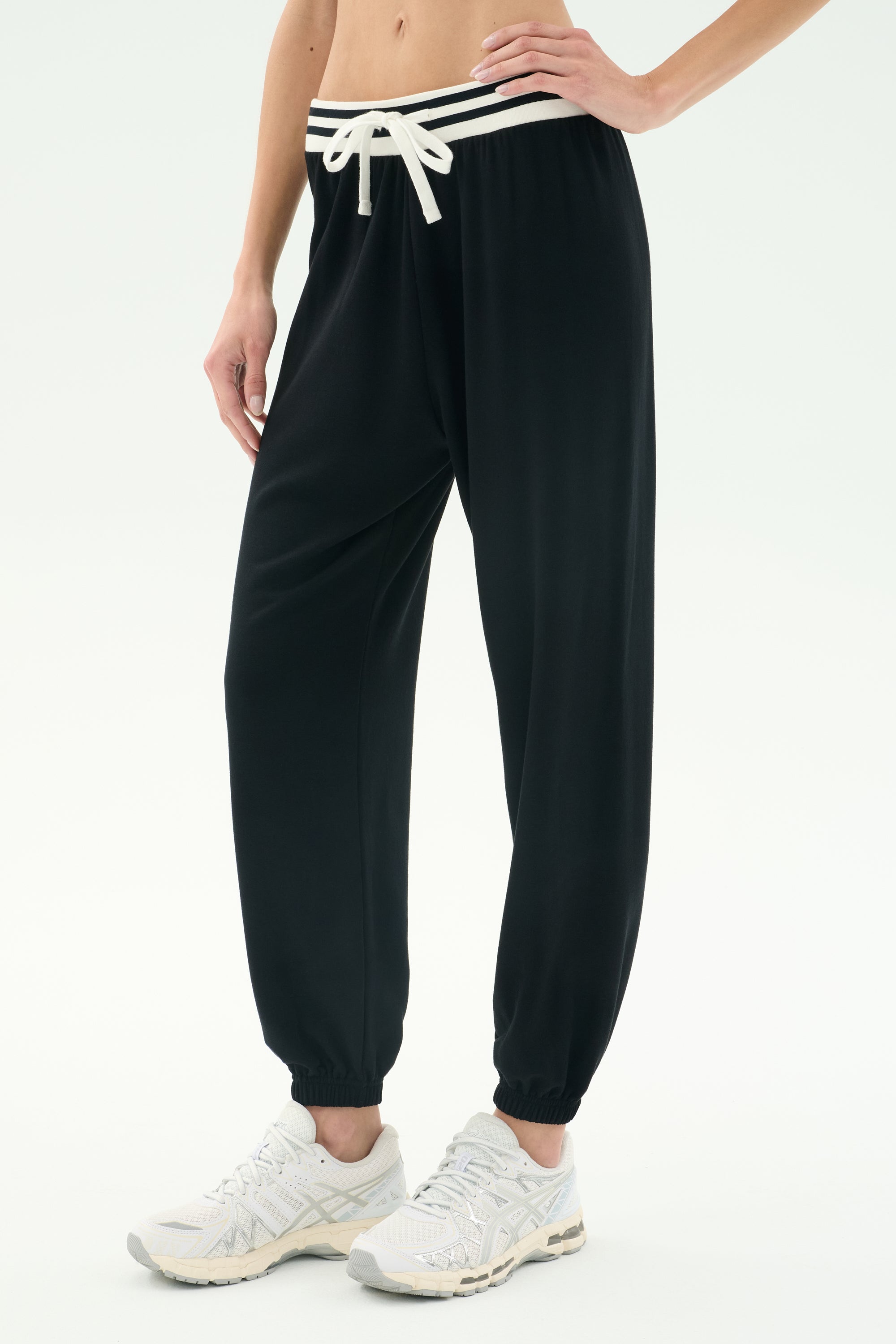 Model wears SPLITS59 Andie Oversized Fleece Sweatpant W/ Stripe in black—featuring soft fleece fabric, a white waistband stripe and drawstring—paired with white athletic shoes and a bare midriff, against a plain light background.