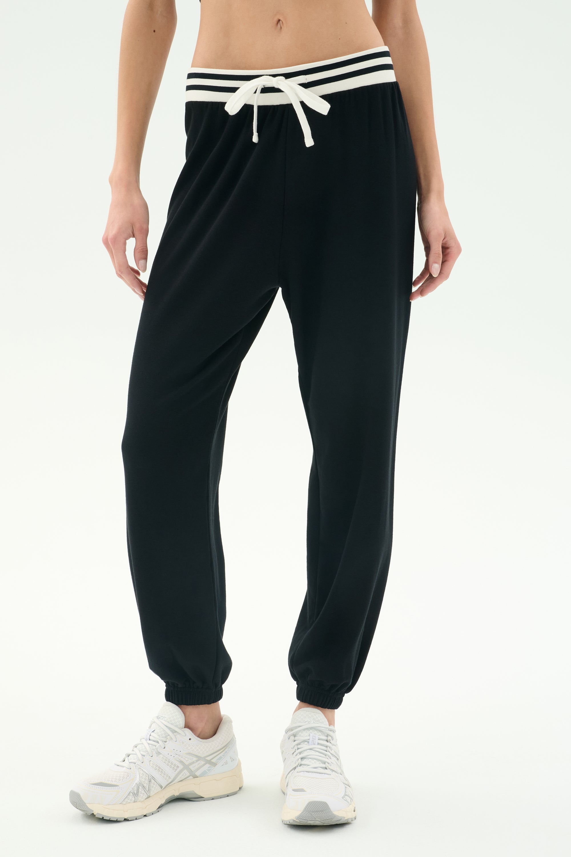 A person wears SPLITS59 Andie Oversized Fleece Sweatpant W/ Stripe in Black with white sneakers, featuring a comfy fit and standing against a plain light background.