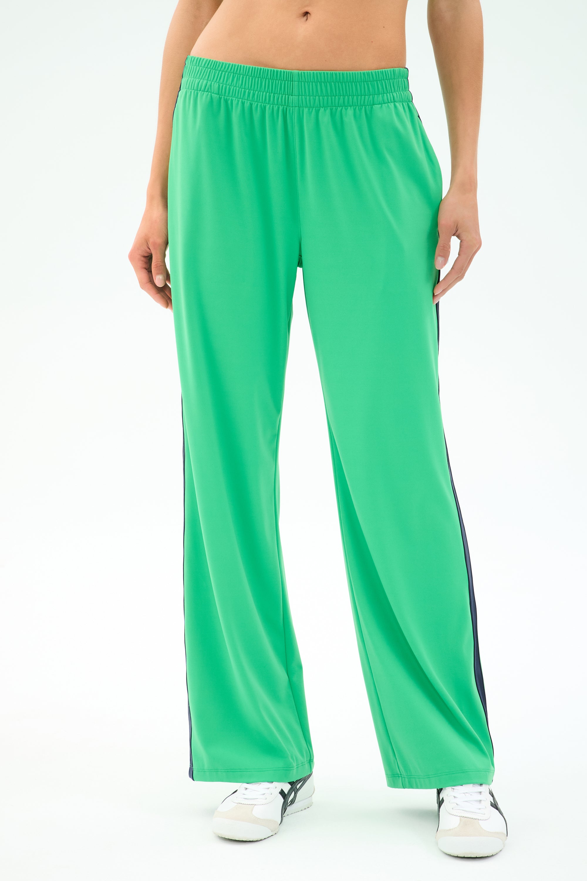 Person wearing SPLITS59 Maxie Rigor Trackpant in Grass/Indigo with black side stripes and white sneakers, standing against a plain background. Only the lower torso and legs are shown.