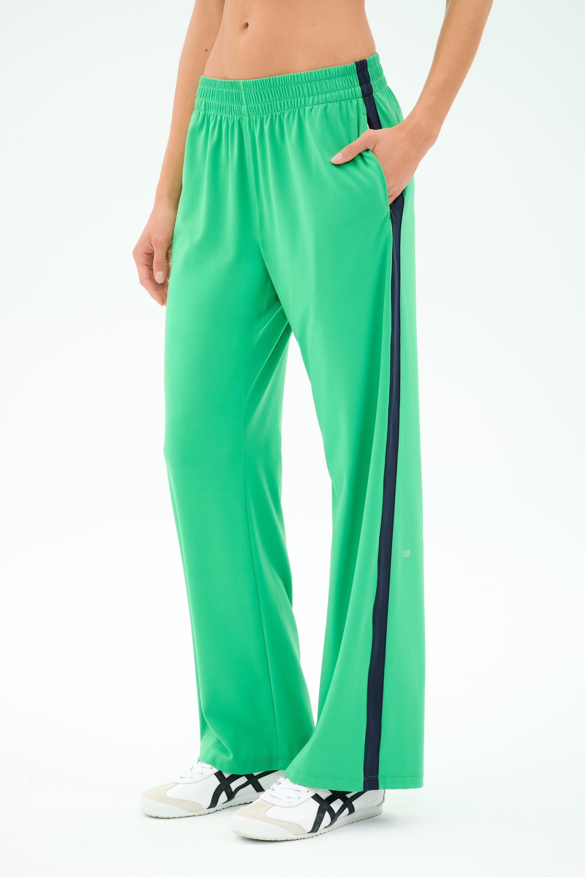 A person wears SPLITS59 Maxie Rigor Trackpant in Grass/Indigo with a black side stripe and white sneakers, standing with one hand in their pocket against a plain backdrop.