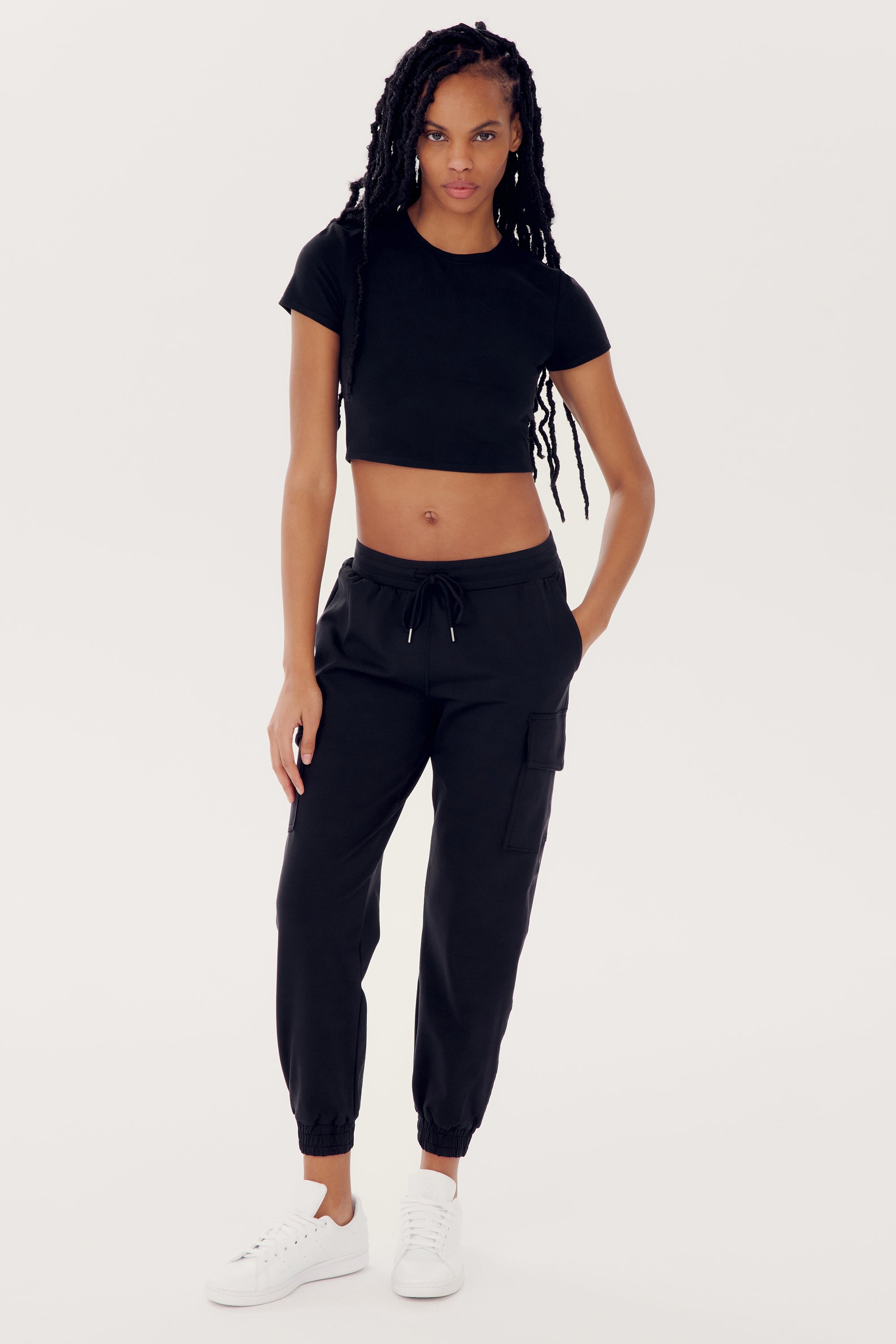Person wearing a black **Airweight S/S Crop - Black** from SPLITS59, black sweatpants, and white sneakers, standing against a plain white background.