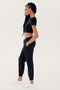 A person with long braids wearing a black SPLITS59 Airweight S/S Crop - Black, black cargo pants, and white sneakers stands in a side view pose against a plain background.