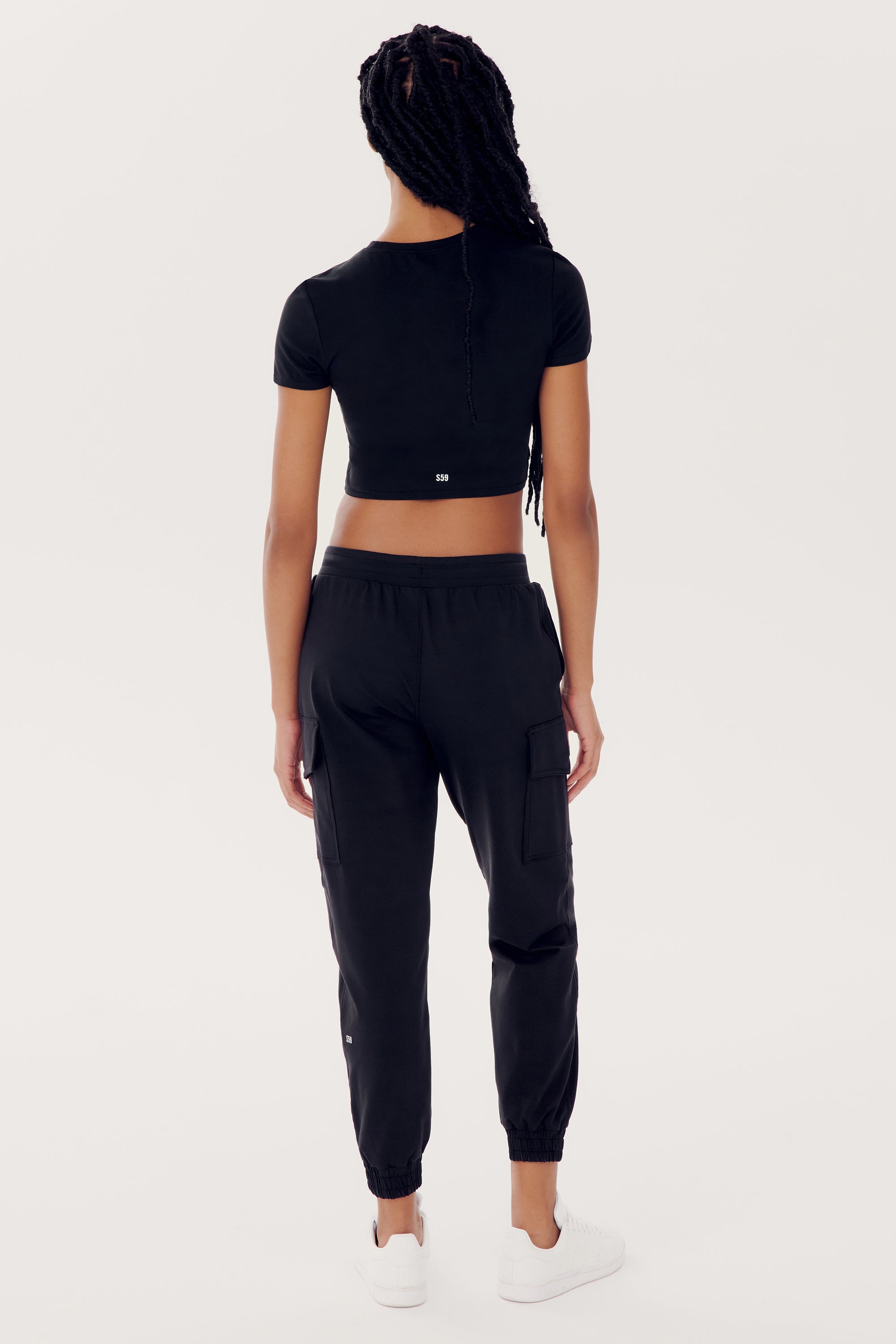 A person with braided hair, wearing a black Airweight S/S Crop by SPLITS59, black jogger pants, and white sneakers, is standing and facing away from the camera.
