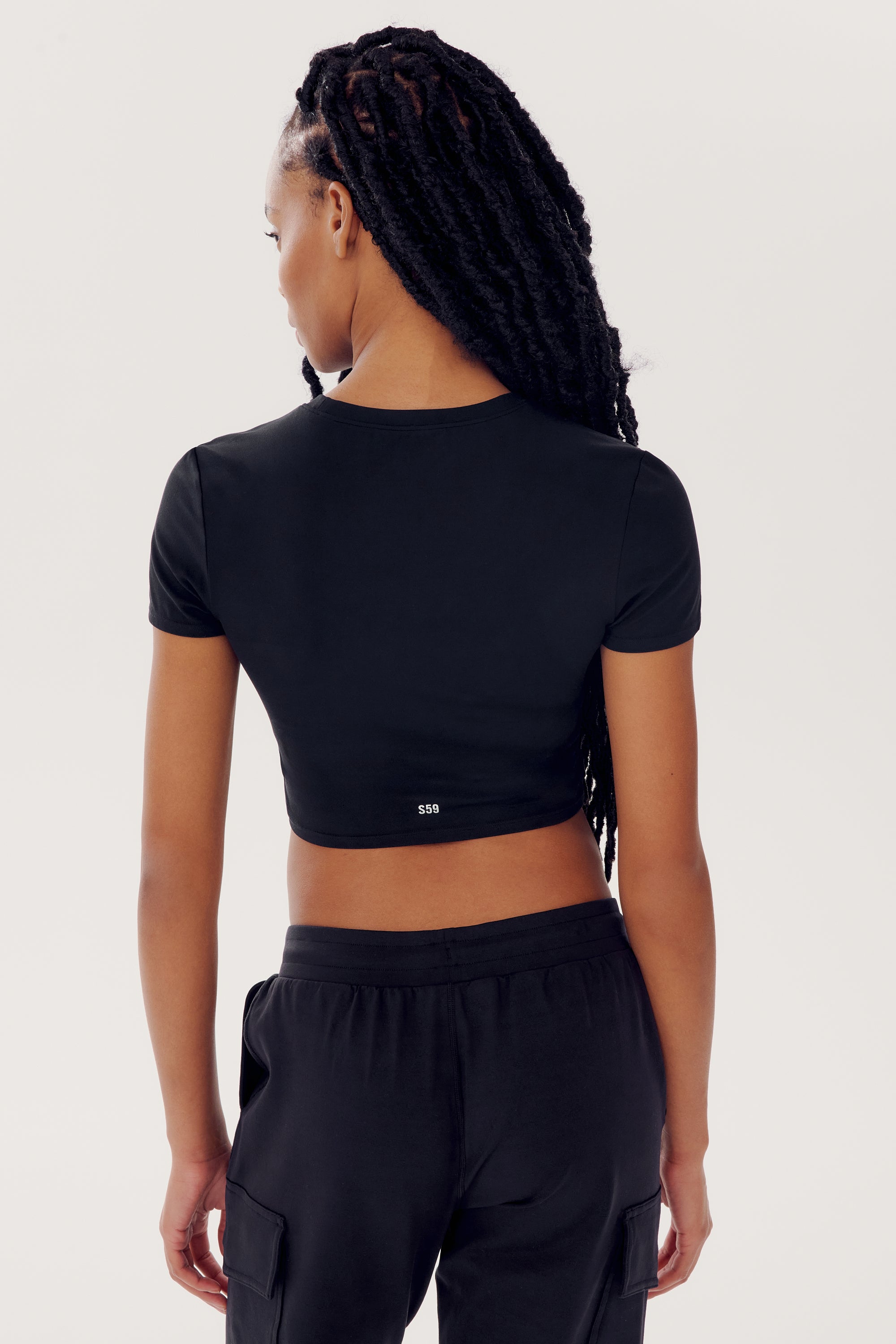 Person with long braided hair wearing a SPLITS59 Airweight S/S Crop - Black and dark pants, viewed from the back against a plain white background.