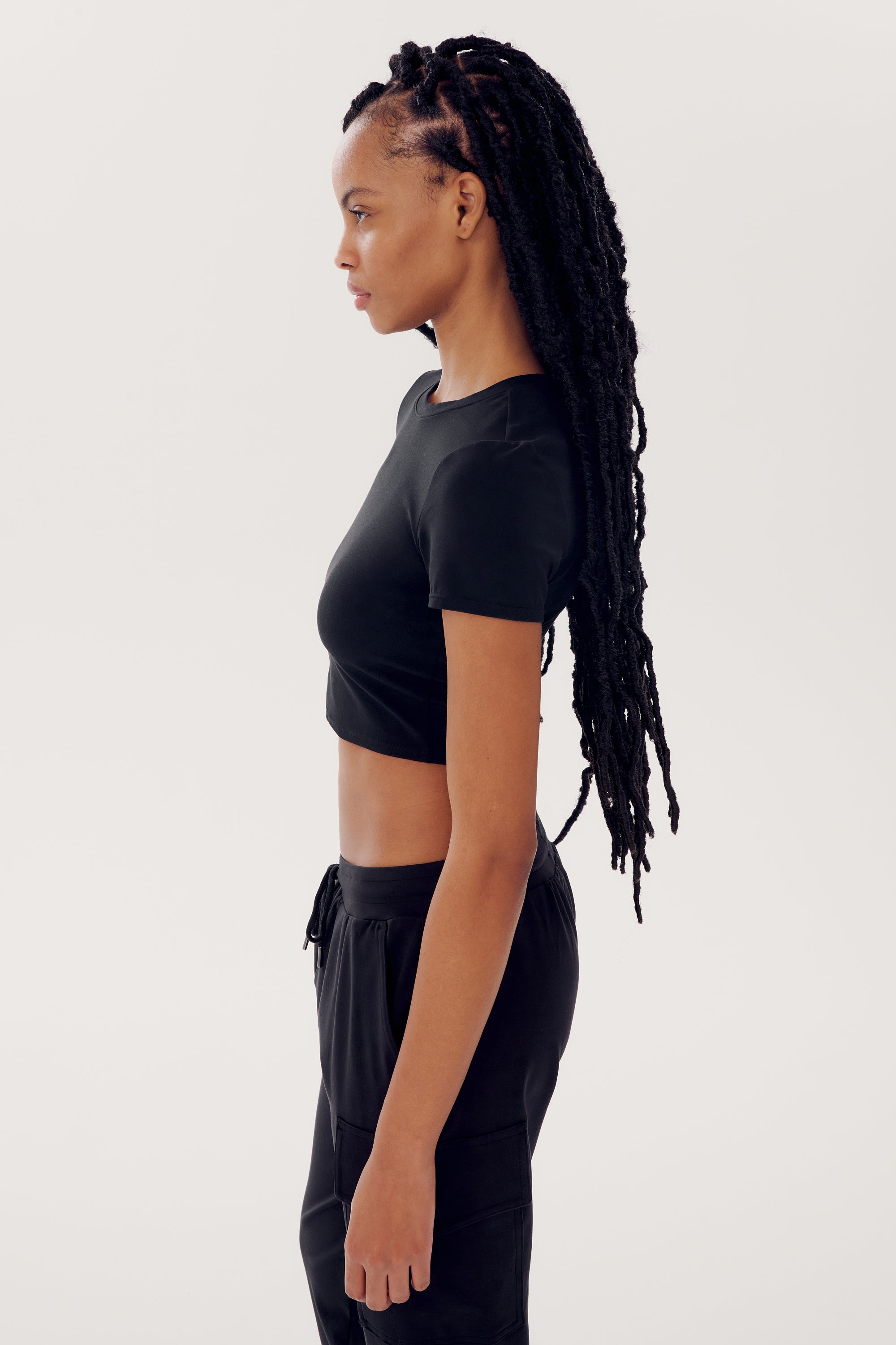 A person with long braids is standing in profile, wearing a black Airweight S/S Crop - Black by SPLITS59 and black pants against a plain white background.