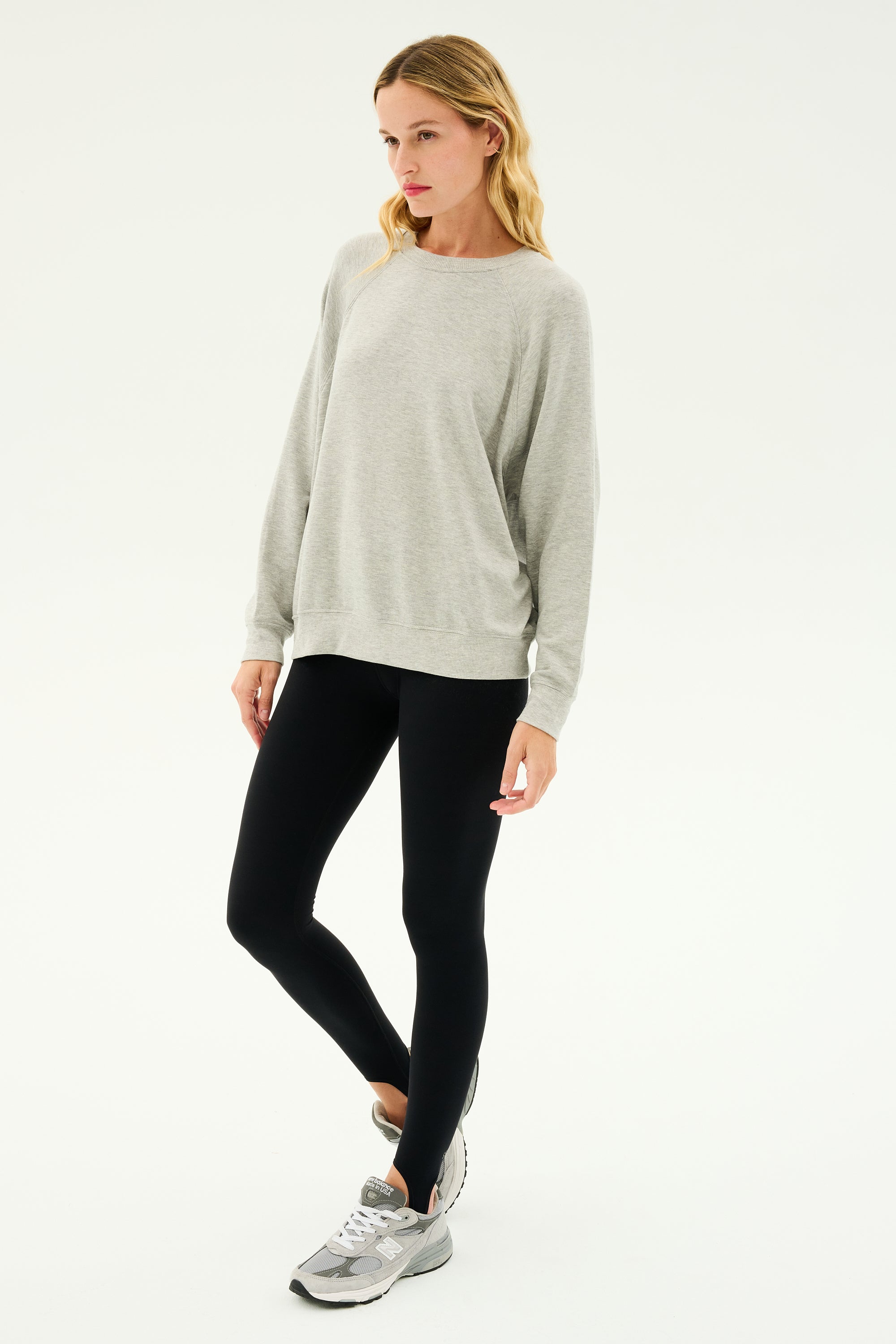 A person wearing an Andie Fleece Sweatshirt in Heather Grey by SPLITS59, black leggings, and grey sneakers stands against a plain white background.