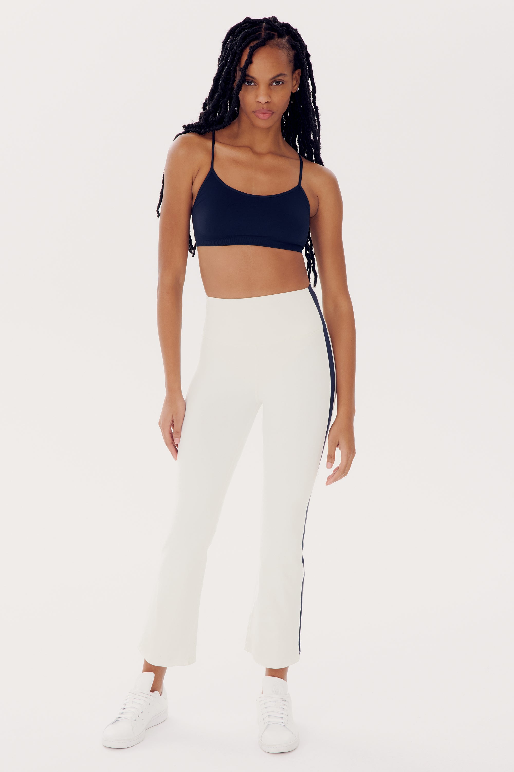 A woman is standing confidently in a black sports bra, high waist SPLITS59 Raquel High Waist Crop - White/Indigo pants with side stripes made of 4-way stretch supplex fabric, and matching white sneakers. She has long braided hair and is posing against a plain white background.