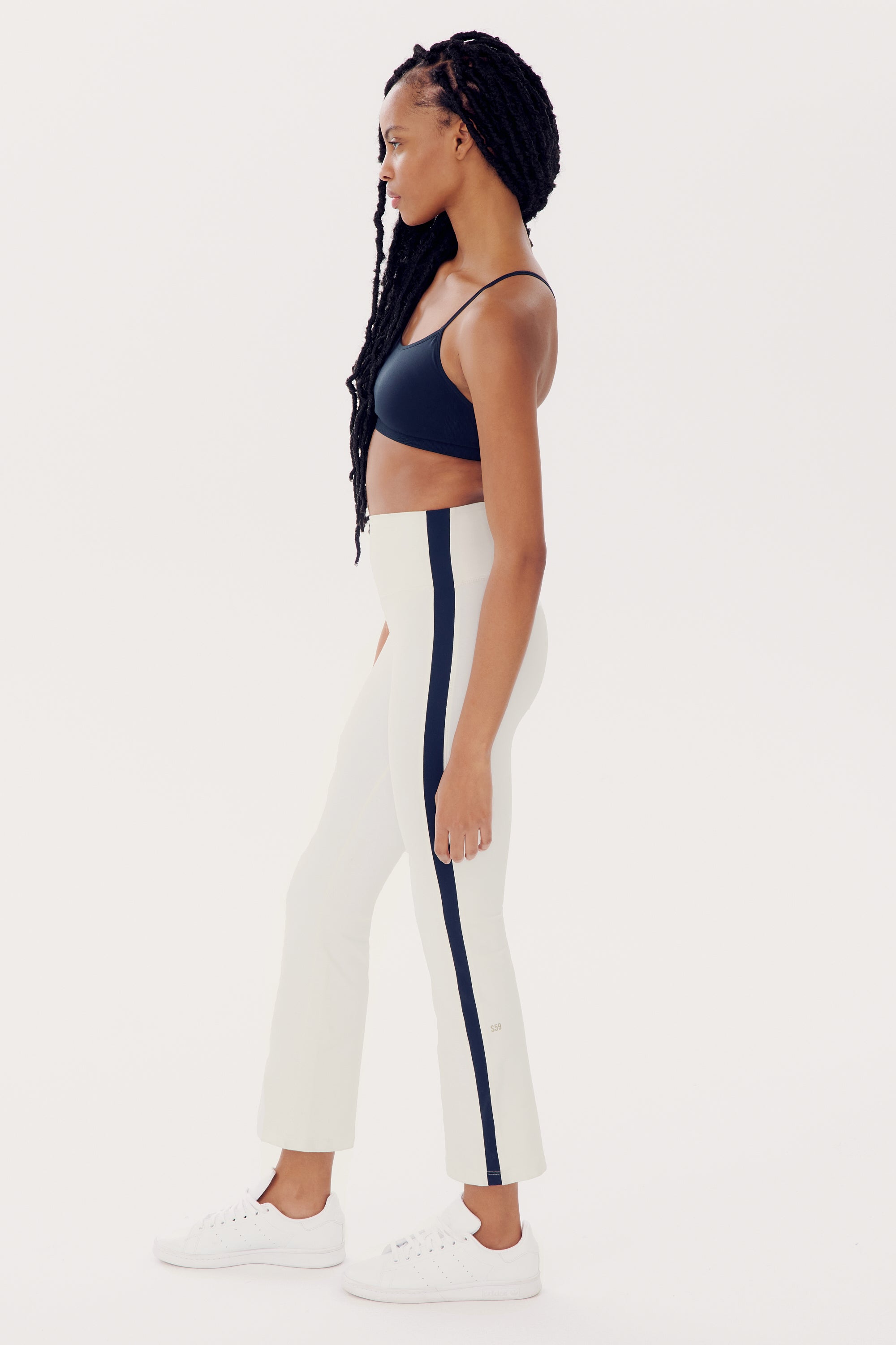 A person with long braids stands in profile, confidently showcasing their SPLITS59 Raquel High Waist Crop - White/Indigo made from 4-way stretch supplex fabric. They wear a black sports bra and white sneakers against a plain white background, epitomizing the essence of an energetic workout.