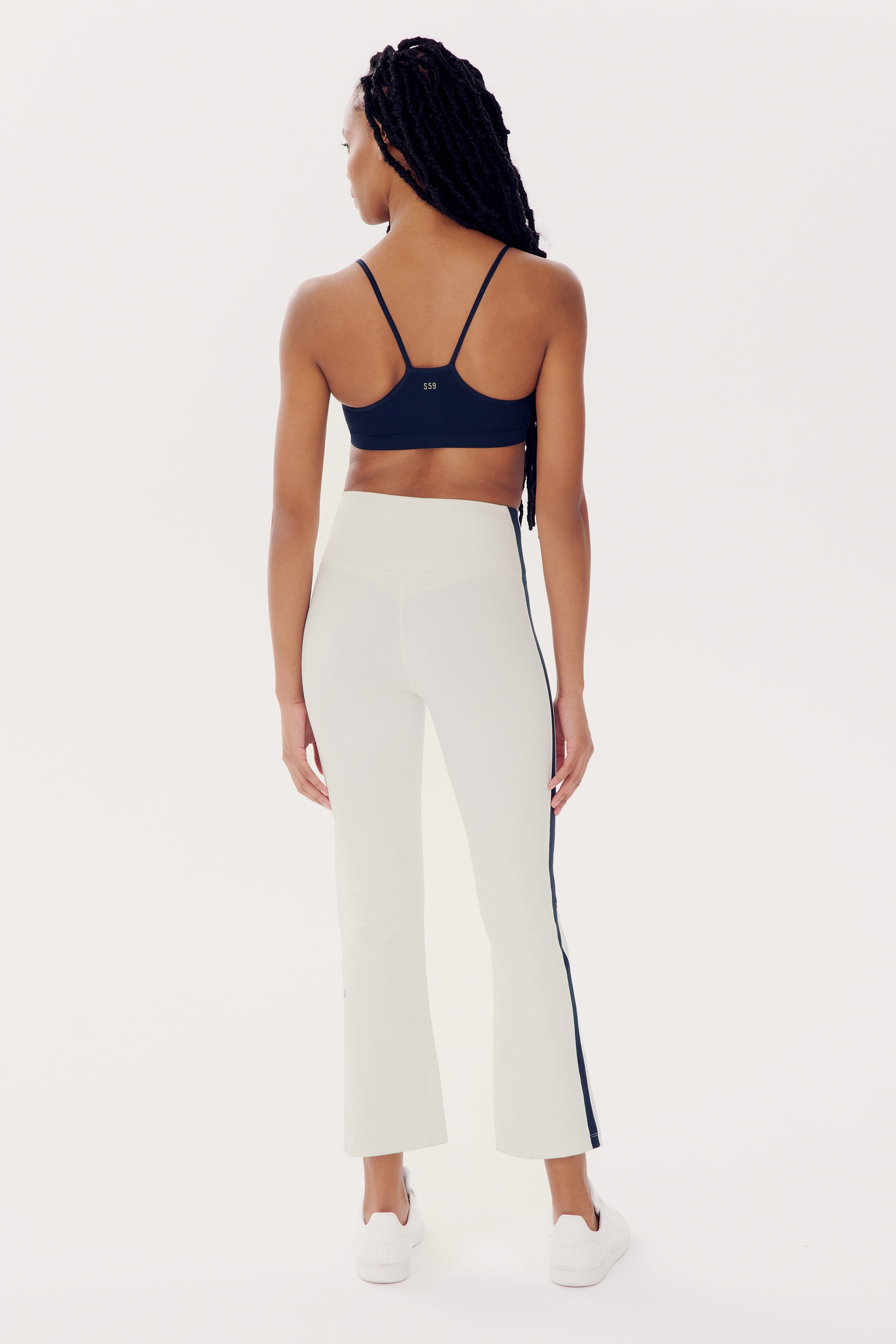 A person with long braided hair is standing, facing away. They radiate confident workout vibes in a navy sports bra and high waist SPLITS59 Raquel High Waist Crop - White/Indigo athletic pants made of 4-way stretch supplex fabric. Their white sneakers complete the look, all set against a plain white background.
