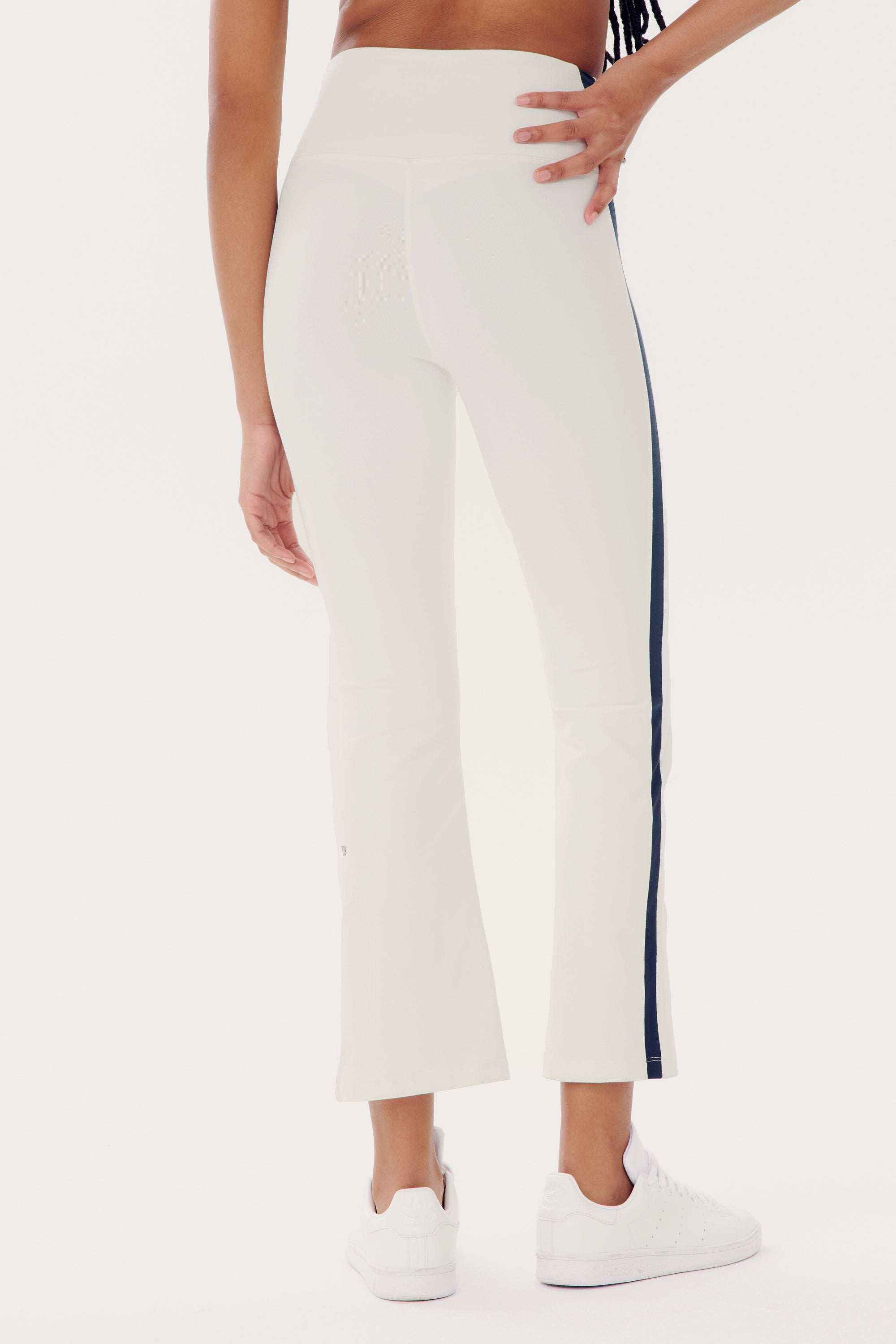 A person is wearing white, high-waist Raquel High Waist Crop - White/Indigo athletic pants from SPLITS59 with a black side stripe and white sneakers. Made from 4-way stretch supplex fabric, these yoga-friendly pants provide flexibility and comfort, as the individual stands with their back facing the camera.