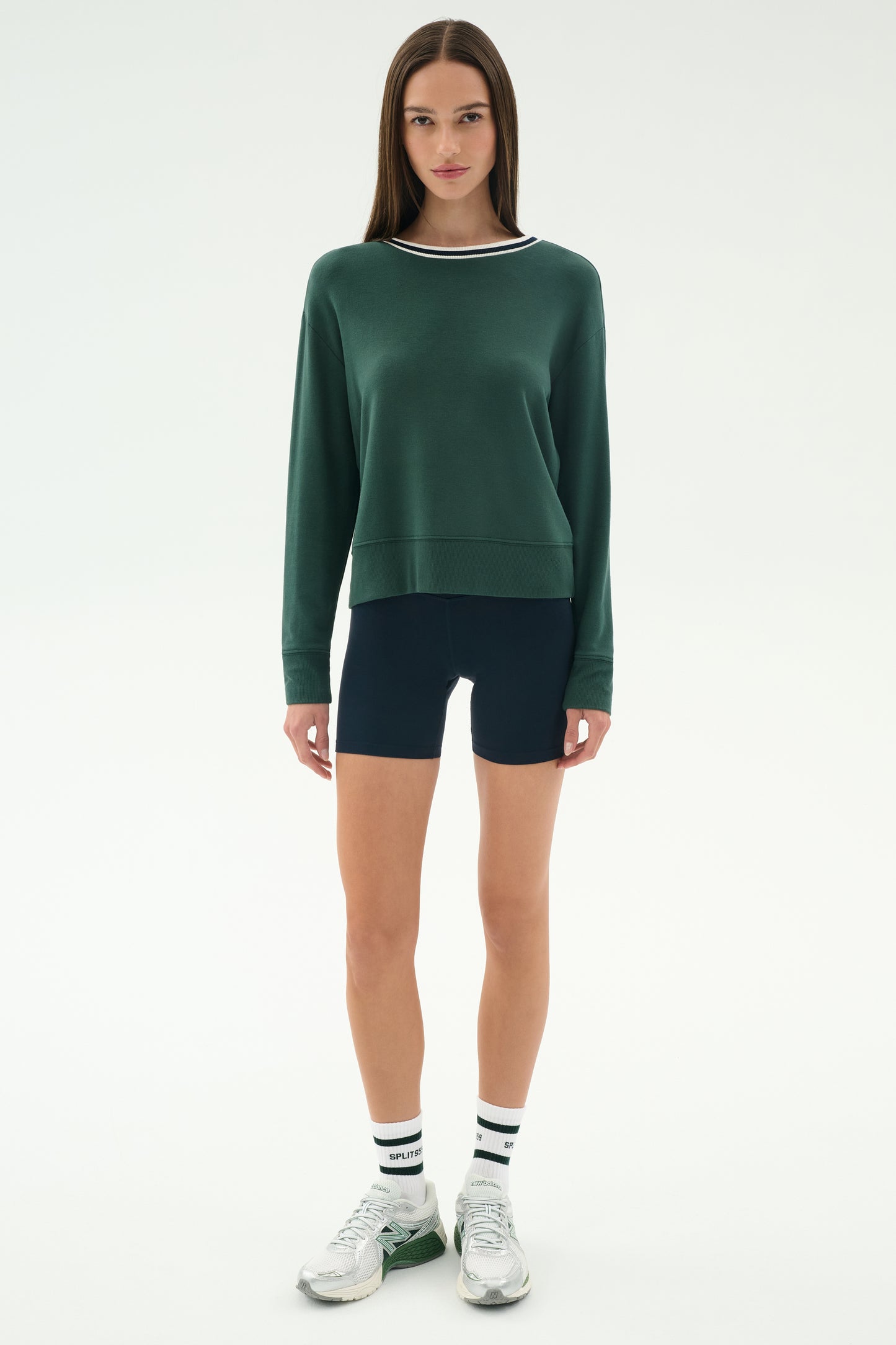 A woman stands against a white background wearing the SPLITS59 Sonja Fleece Sweatshirt w/ Stripe in Military, paired with black bike shorts, white socks with black stripes, and white sneakers.