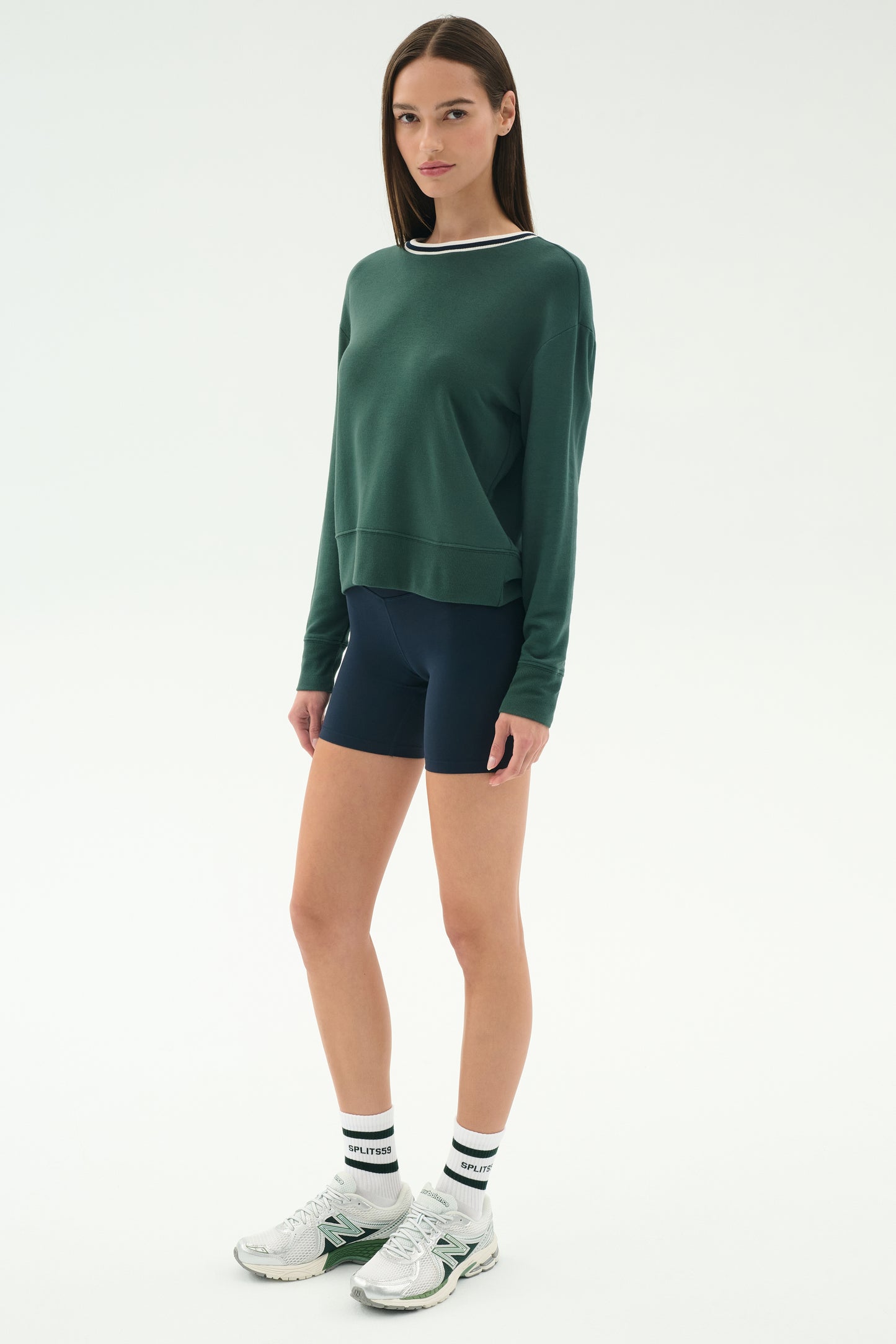 A woman stands facing forward wearing the SPLITS59 Sonja Fleece Sweatshirt with Stripe in Military, navy spandex bike shorts, white striped socks, and silver sneakers against a plain light background.