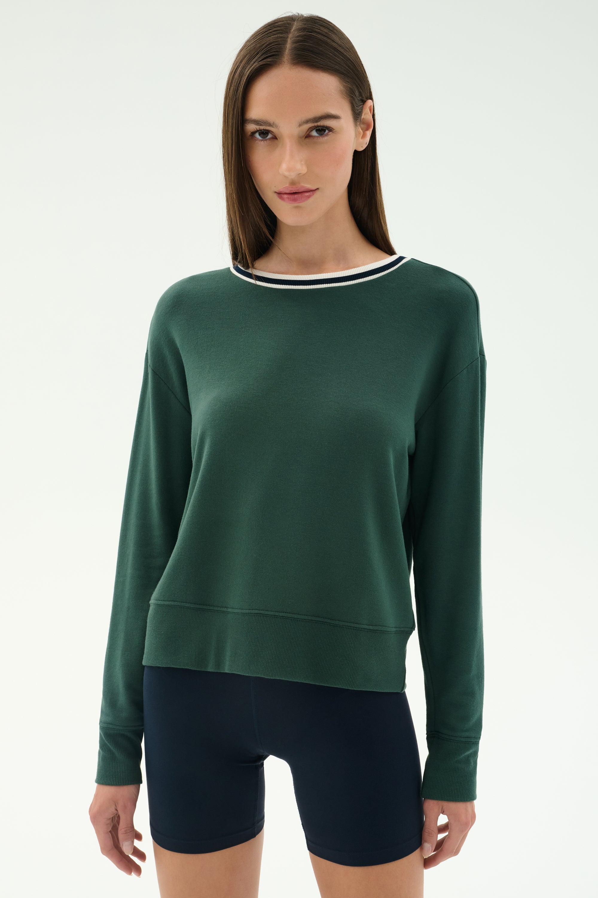 A woman faces forward in a SPLITS59 Sonja Fleece Sweatshirt W/ Stripe in military green, featuring soft modal fabric and a white-trimmed collar, paired with navy cycling shorts against a plain light background.
