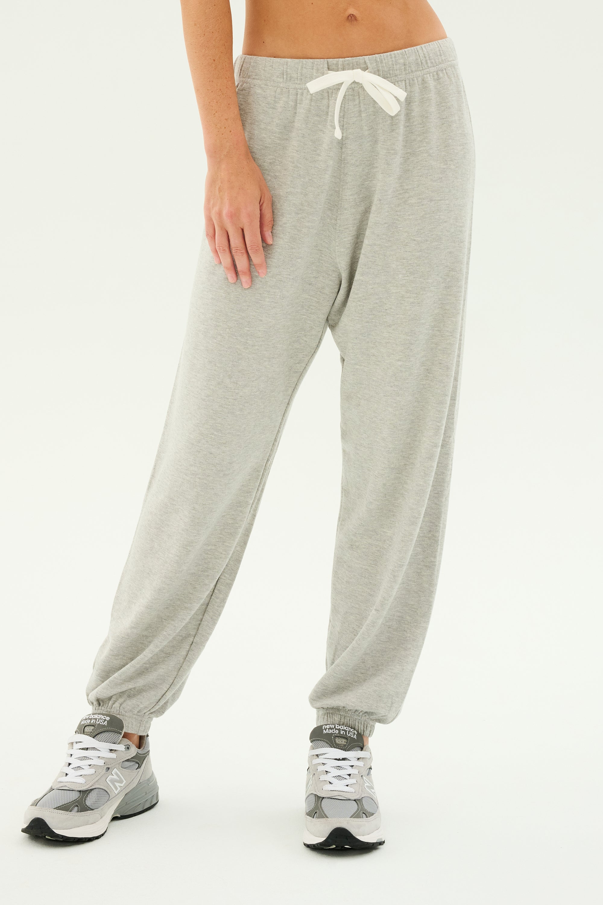 A person wearing the Andie Oversized Fleece Sweatpant in Heather Grey by SPLITS59 and gray sneakers stands on a plain background. The sweatpants, perfect for cool weather workouts or relaxing at home, feature an elastic waistband with a white drawstring. Only the lower half of the body is visible.