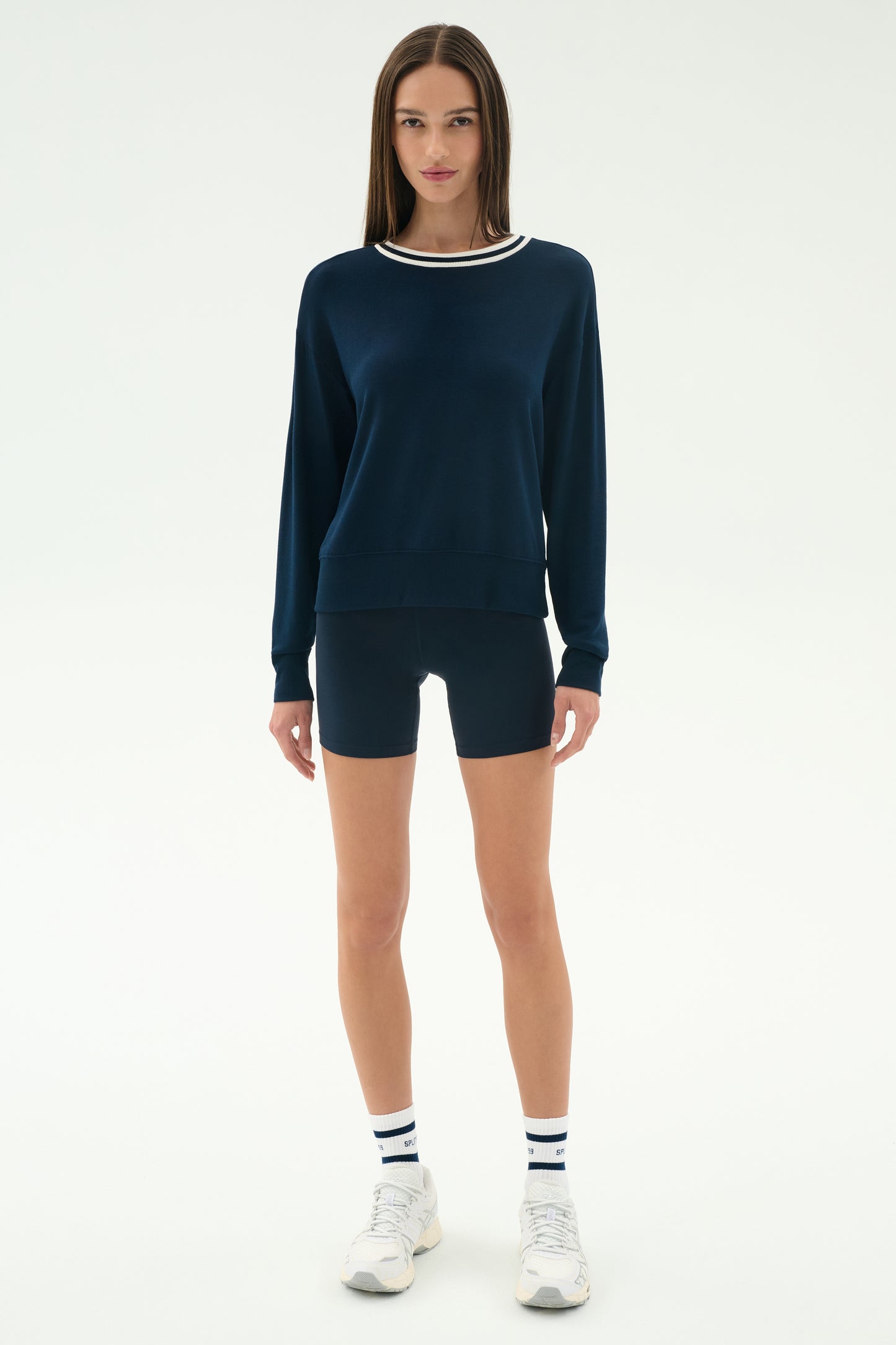 A woman stands facing forward in the SPLITS59 Sonja Fleece Sweatshirt W/ Stripe in Indigo, paired with matching navy shorts, striped white athletic socks, and white sneakers against a plain white background.