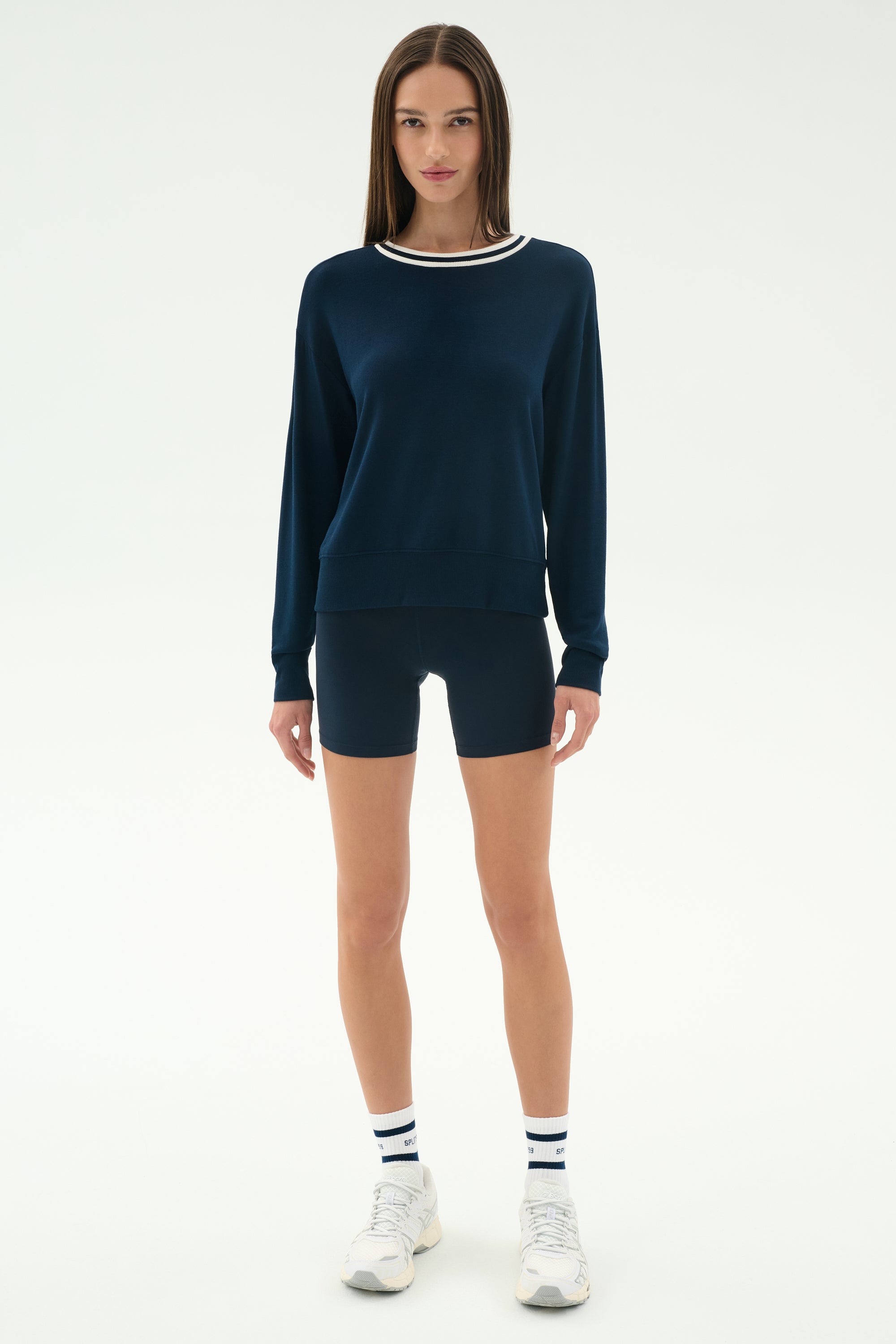 A woman stands facing forward in the SPLITS59 Sonja Fleece Sweatshirt W/ Stripe in Indigo, paired with matching navy shorts, striped white athletic socks, and white sneakers against a plain white background.