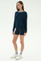 A woman stands against a plain background wearing the SPLITS59 Sonja Fleece Sweatshirt W/ Stripe in Indigo, paired with navy bike shorts, white sneakers, and white socks.