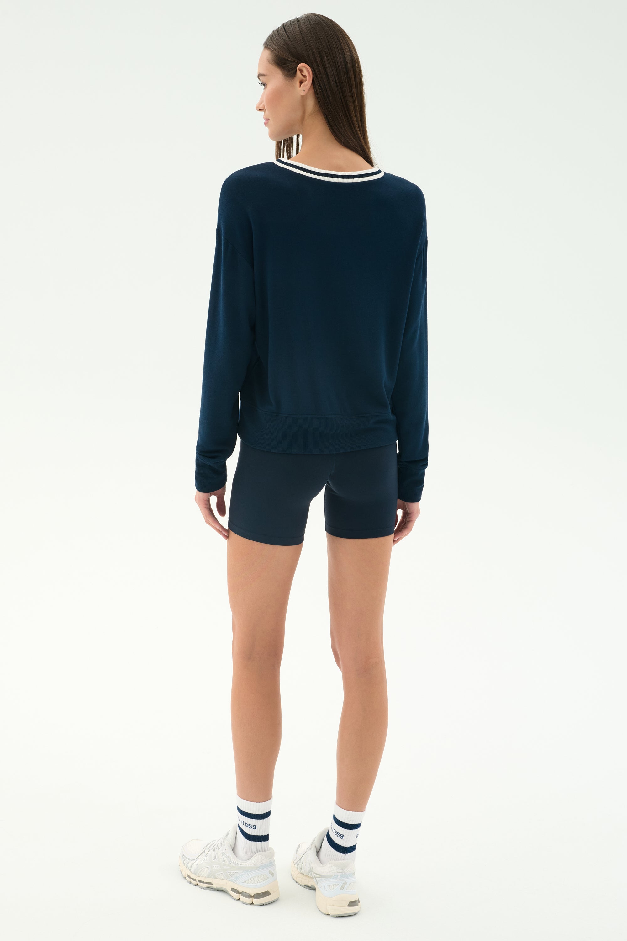 A woman faces away, wearing the SPLITS59 Sonja Fleece Sweatshirt w/ Stripe in indigo, navy shorts, white crew socks with stripes, and white sneakers against a white background.