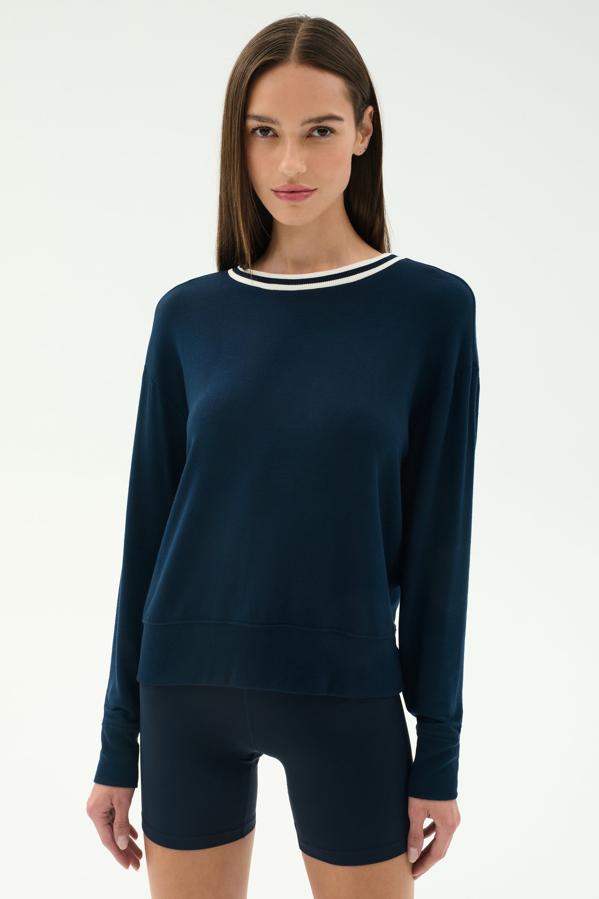 A woman with straight brown hair wears the SPLITS59 Sonja Fleece Sweatshirt W/ Stripe in indigo, paired with matching shorts, standing against a plain light background.