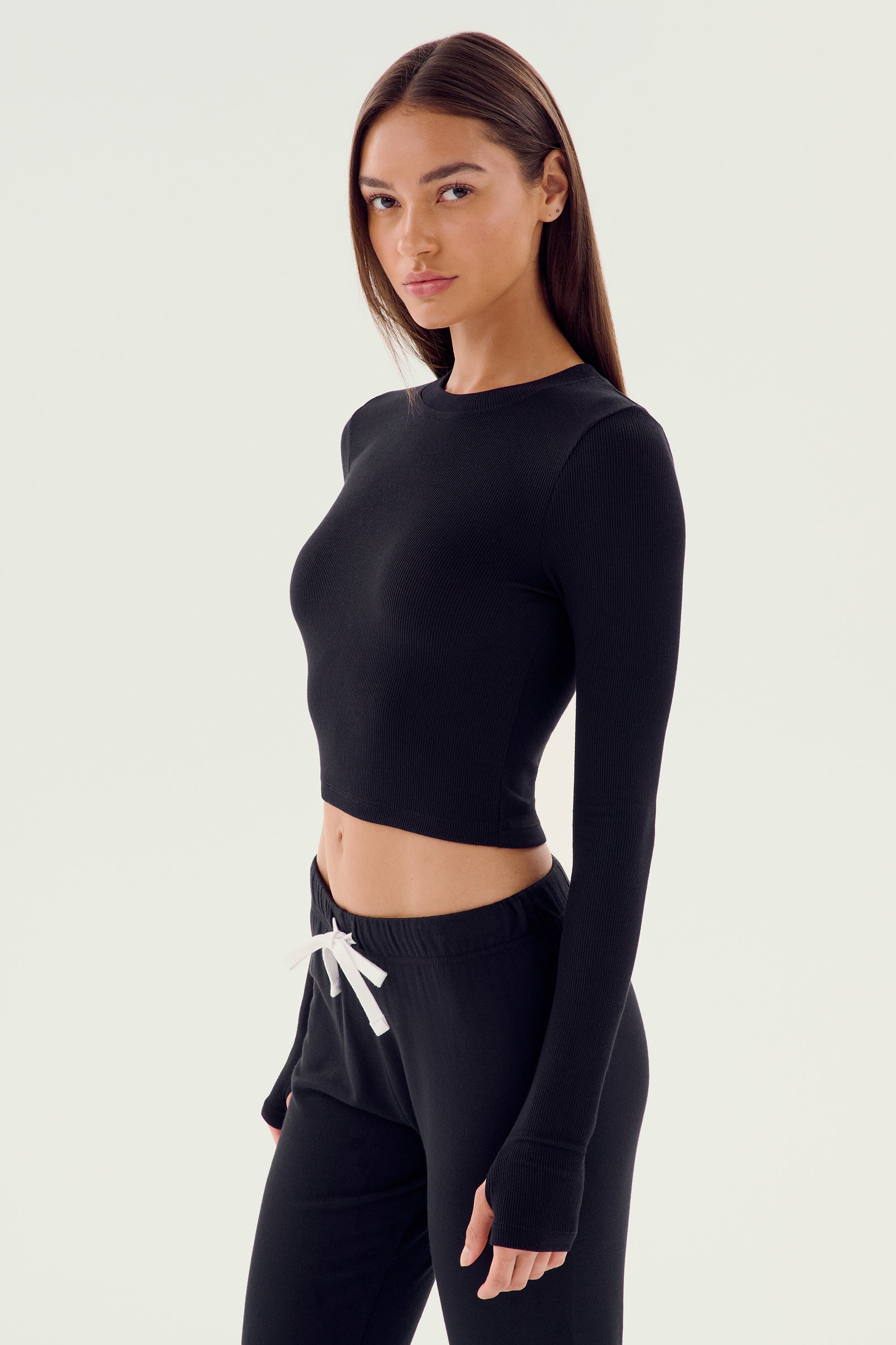 A woman stands against a plain background, wearing the SPLITS59 Louise Rib Long Sleeve Crop - Black and black pants, perfect for gym workouts.