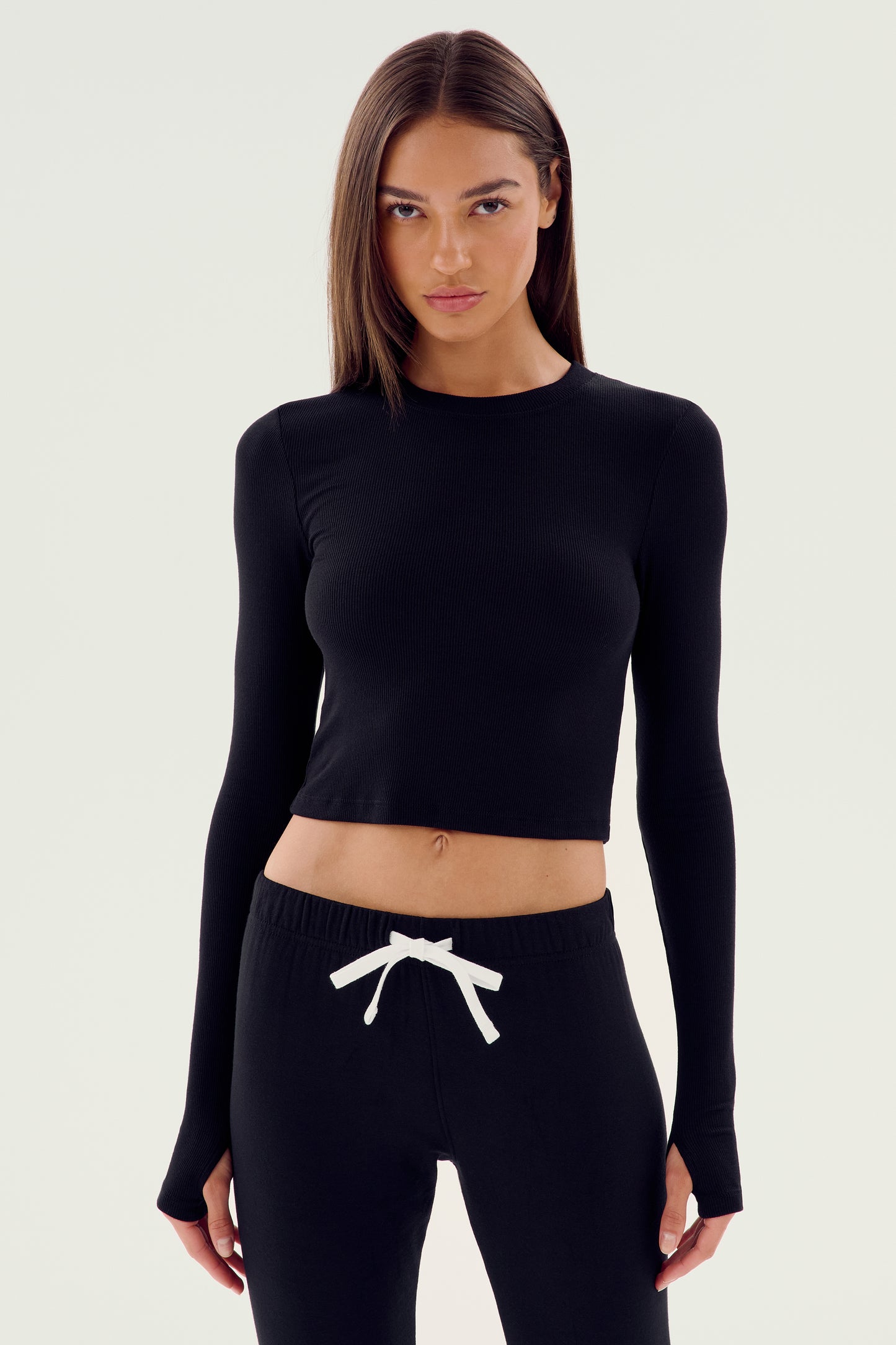 A person with long hair is posing against a plain background, clad in the SPLITS59 Louise Rib Long Sleeve Crop - Black and matching black drawstring pants.
