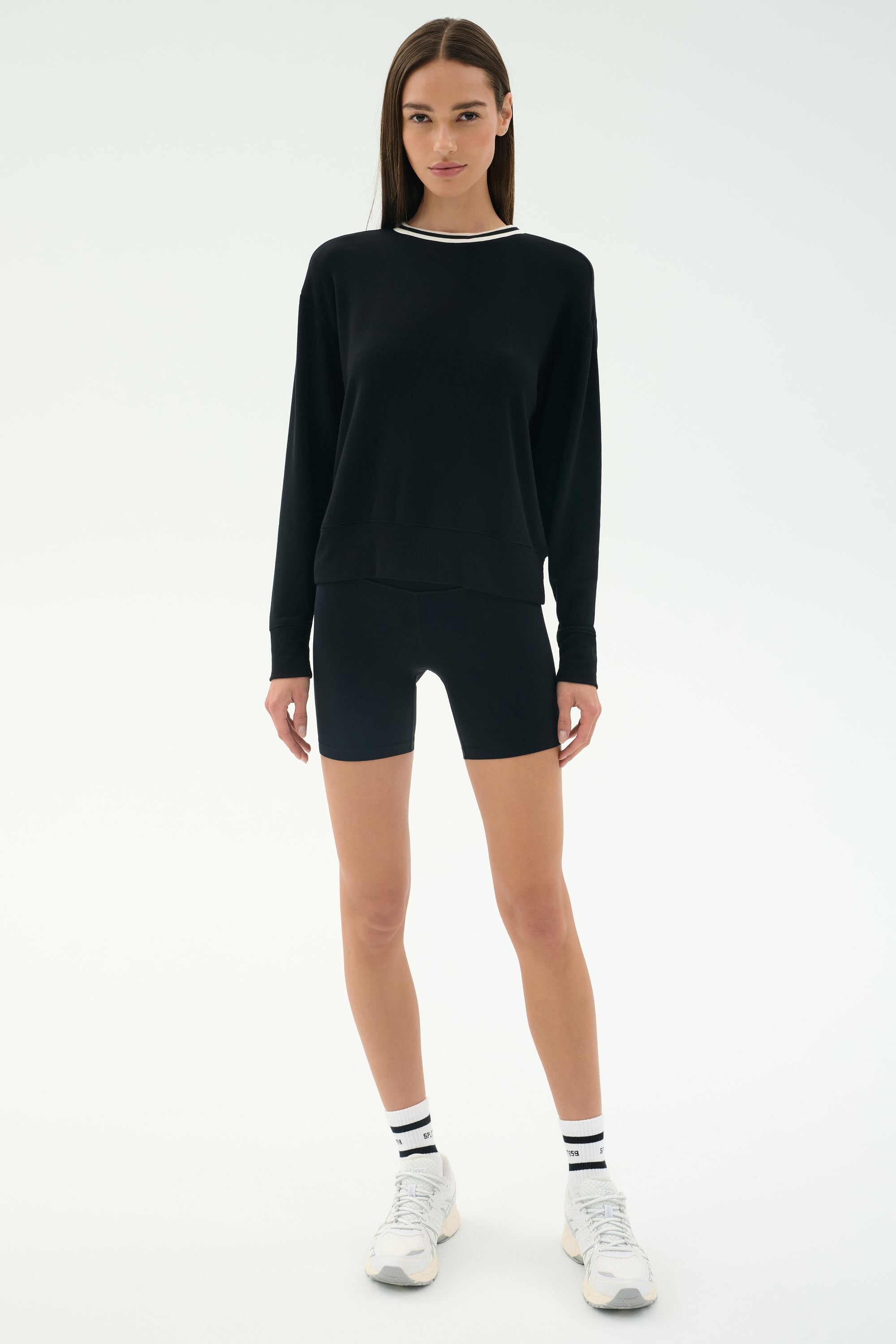 A woman faces forward on a plain light background wearing the SPLITS59 Sonja Fleece Sweatshirt W/ Stripe, black fitted shorts, white sneakers, and white socks with black stripes.