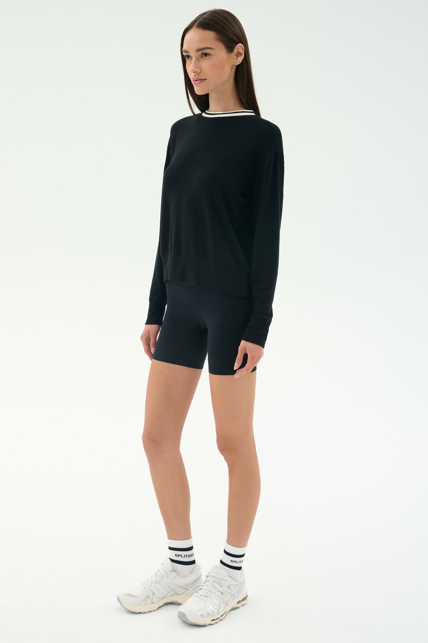 A person stands facing slightly left wearing the SPLITS59 Sonja Fleece Sweatshirt W/ Stripe in black, paired with black cycling shorts, white crew socks, and white athletic shoes against a plain white background.