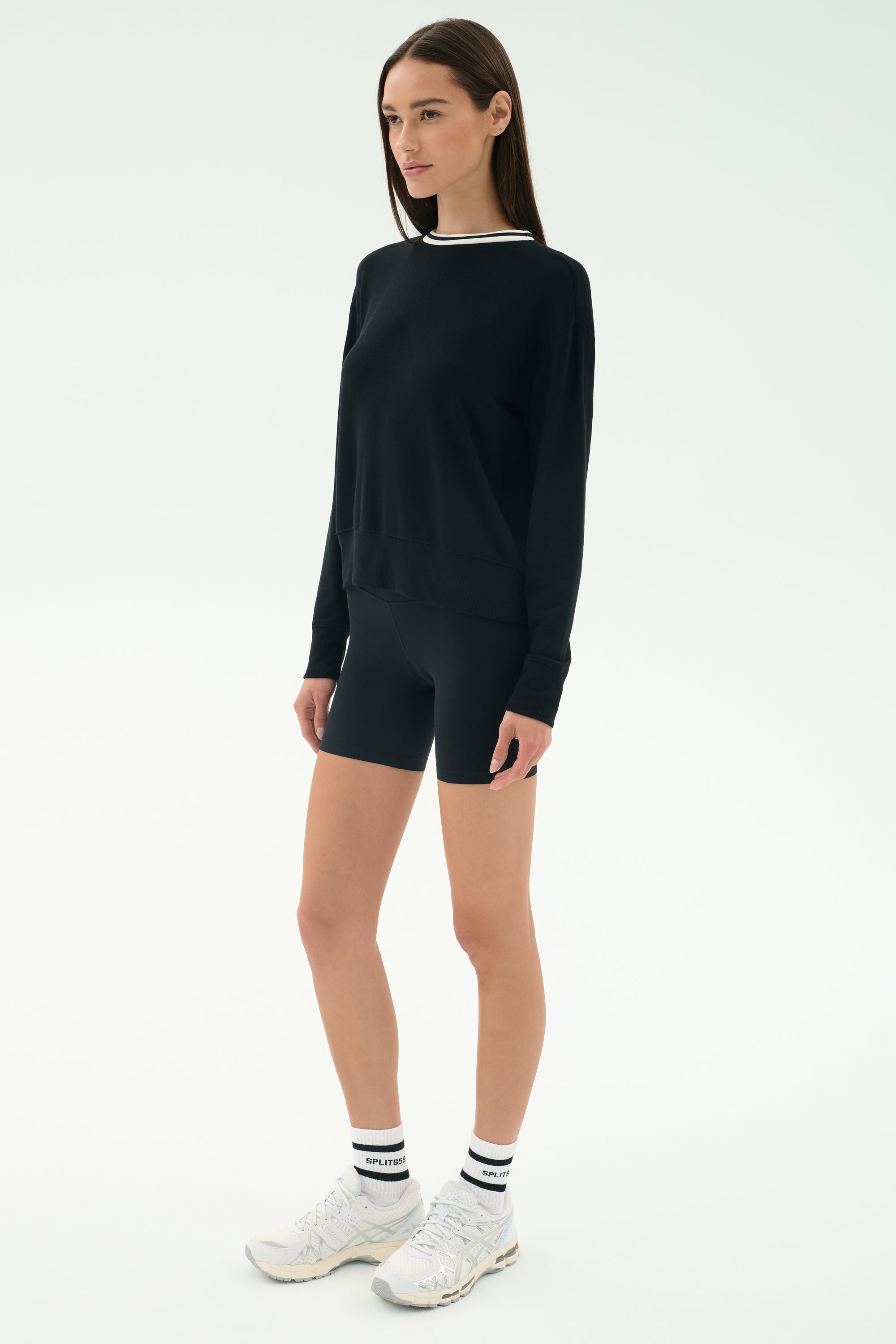 A person stands facing slightly left wearing the SPLITS59 Sonja Fleece Sweatshirt W/ Stripe in black, paired with black cycling shorts, white crew socks, and white athletic shoes against a plain white background.