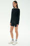 A person stands facing slightly left wearing the SPLITS59 Sonja Fleece Sweatshirt W/ Stripe in black, paired with black cycling shorts, white crew socks, and white athletic shoes against a plain white background.