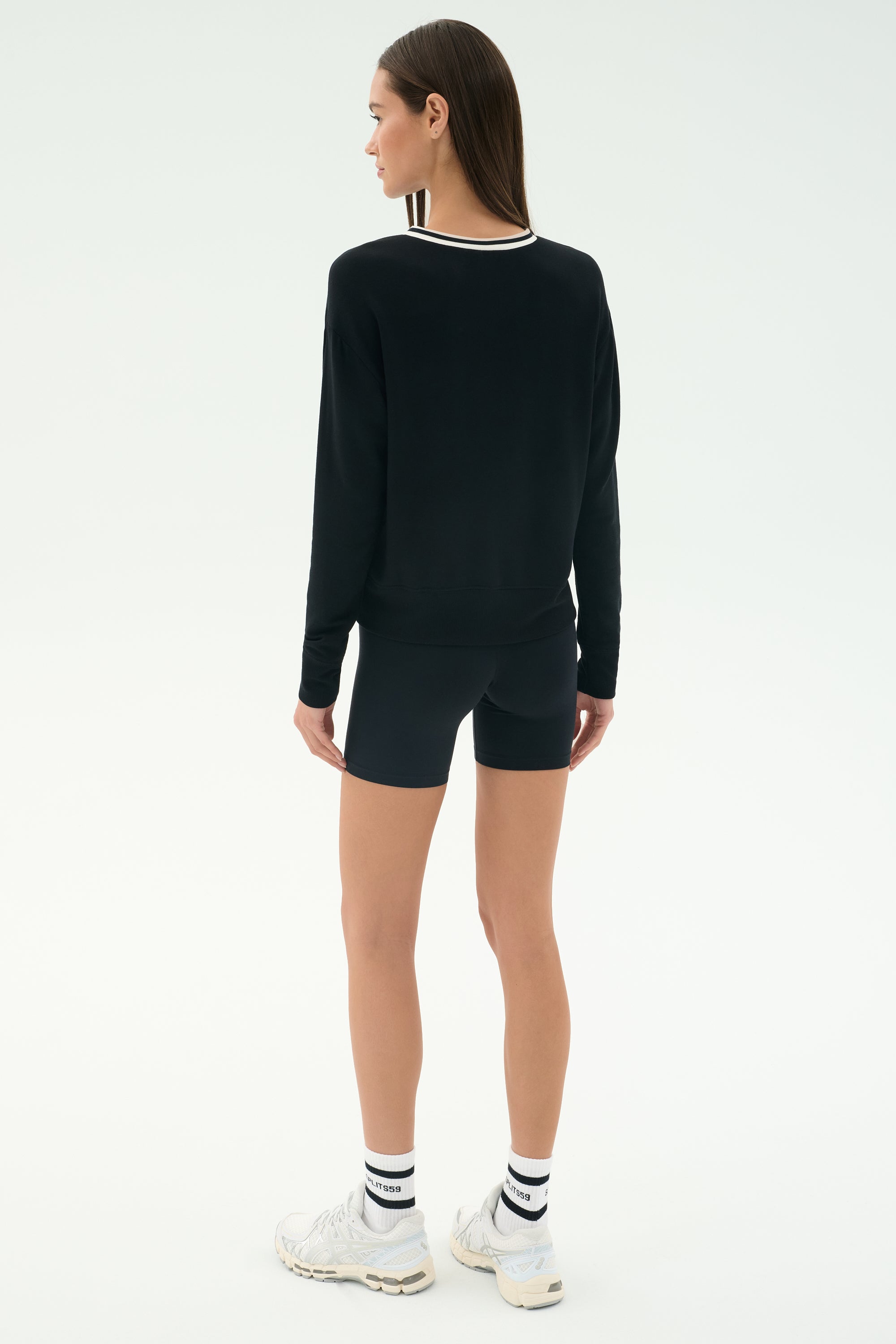 A woman stands with her back to the camera, wearing the SPLITS59 Sonja Fleece Sweatshirt W/ Stripe in black, paired with black shorts, white socks, and white sneakers against a plain light background.
