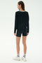 A woman stands with her back to the camera, wearing the SPLITS59 Sonja Fleece Sweatshirt W/ Stripe in black, paired with black shorts, white socks, and white sneakers against a plain light background.