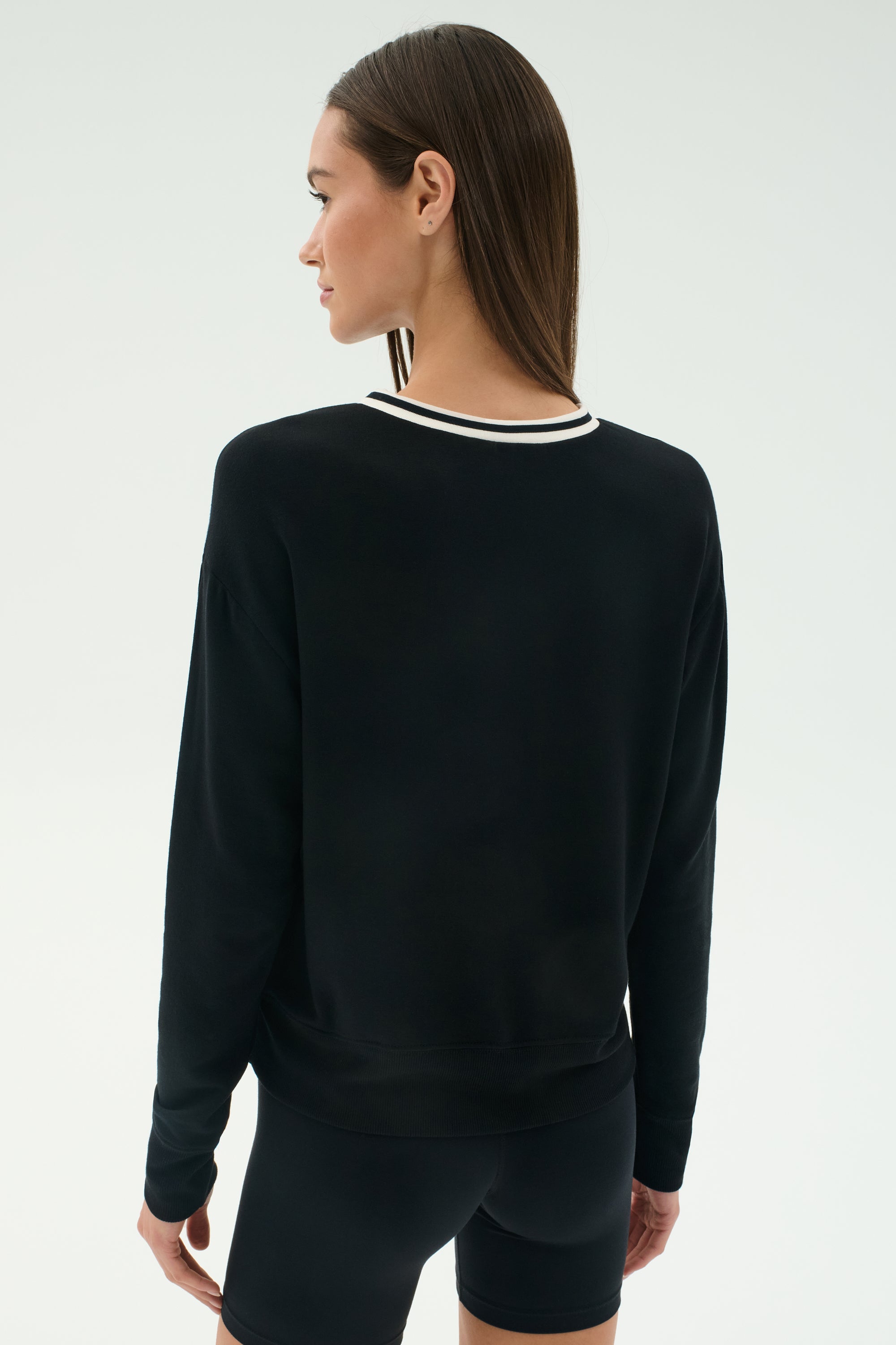 A woman with straight brown hair is seen from the back wearing the SPLITS59 Sonja Fleece Sweatshirt W/ Stripe in black, paired with black modal shorts, against a plain light background.