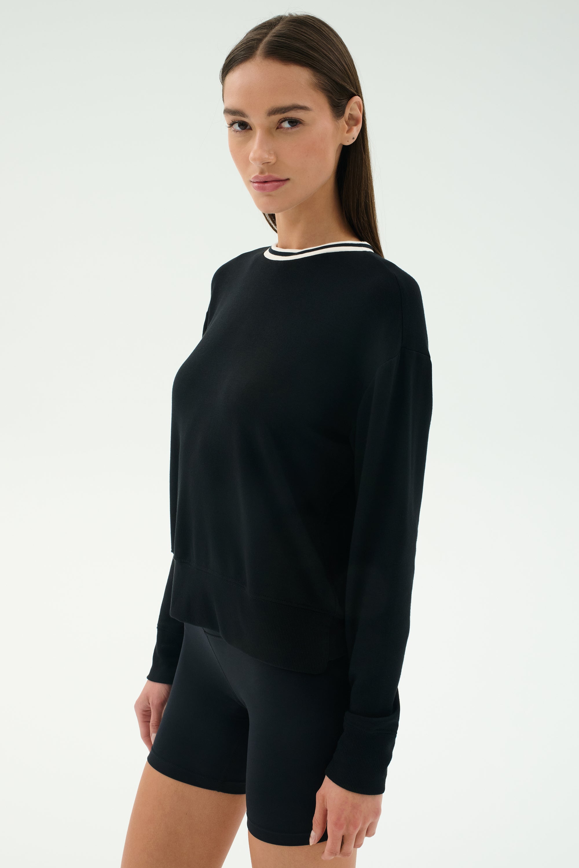 A woman stands against a plain background, wearing the SPLITS59 Sonja Fleece Sweatshirt W/ Stripe in black, looking towards the camera.