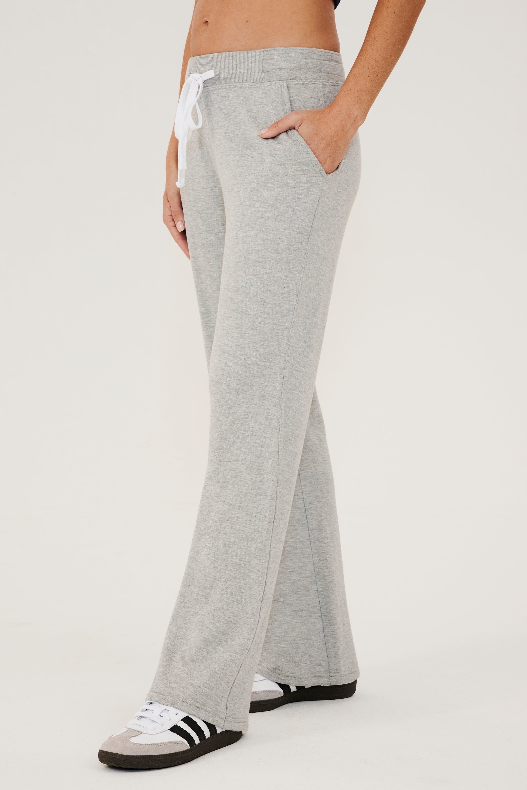 Front side view of woman wearing a light gray high rise wide leg relaxed fit sweatpant with side pockets and white drawstring. Paired with white and grey shoes with black stripes.