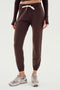 A person is wearing the Sonja Fleece Sweatpant - Dark Chocolate by SPLITS59, featuring a white drawstring, paired with beige sneakers. Perfect for cool weather workouts, the minimalistic backdrop is plain and white.