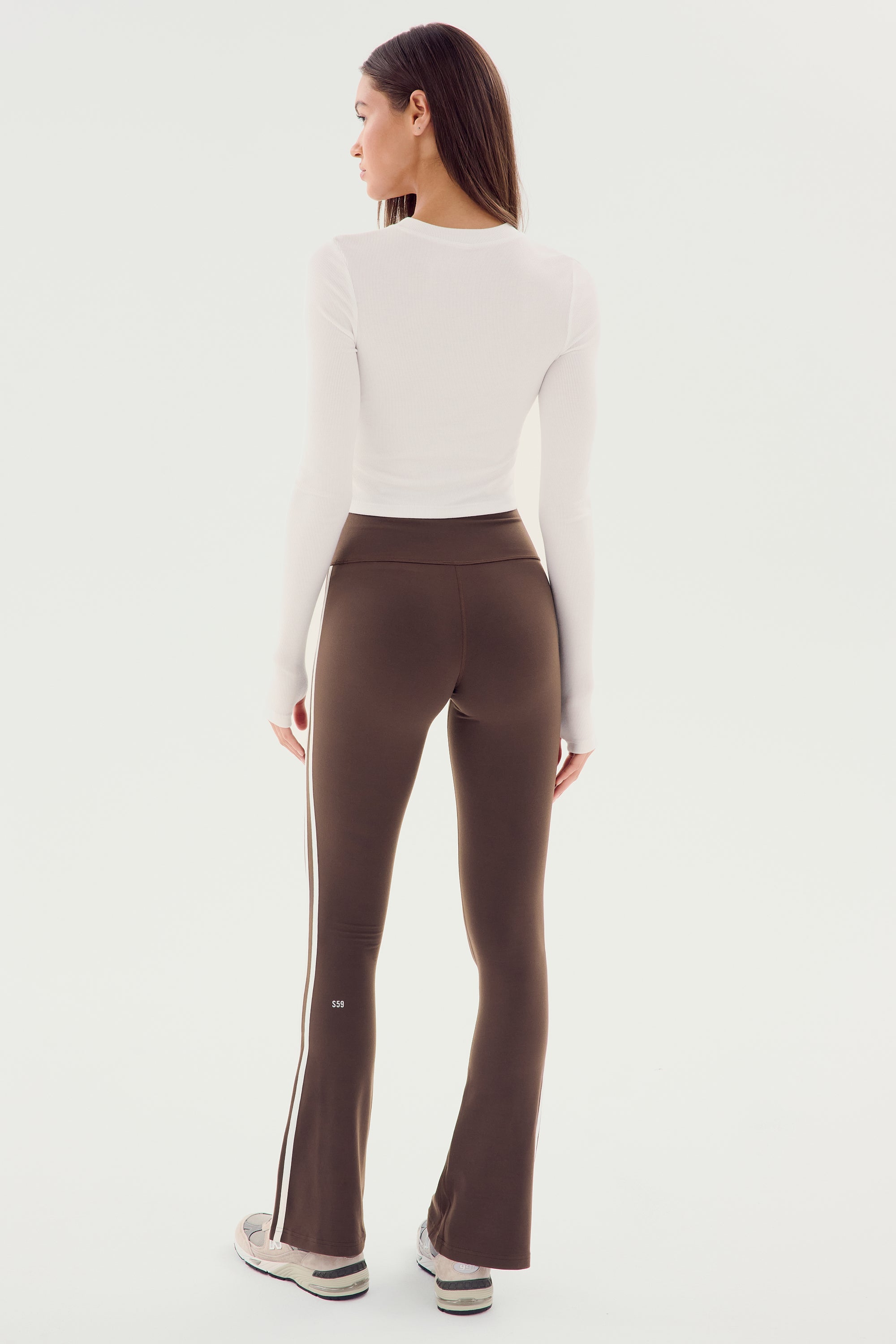 A person is standing and facing away, wearing a white SPLITS59 Louise Rib Long Sleeve Crop, brown flare leggings with a side stripe, and sneakers.