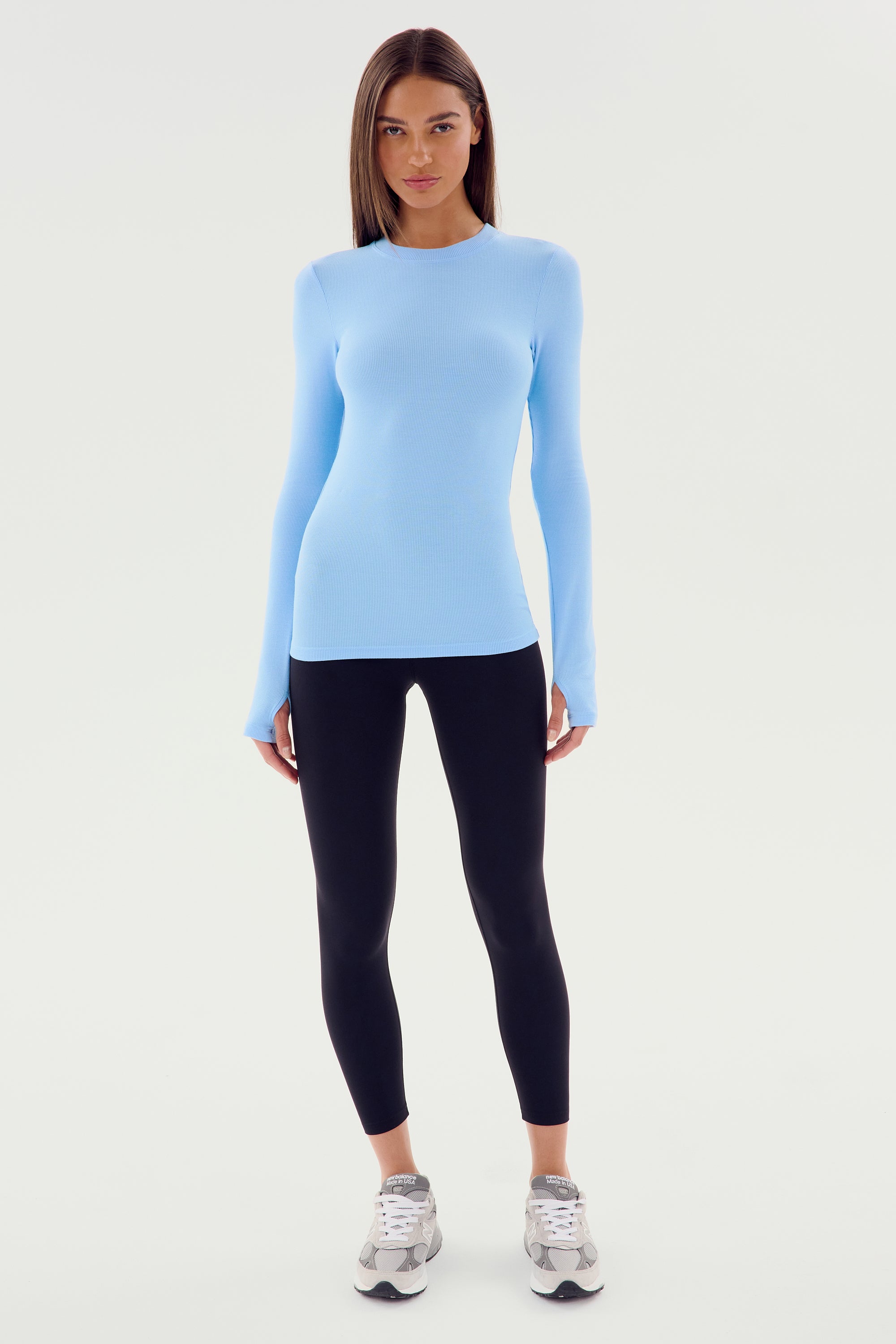 A woman stands facing forward, wearing a SPLITS59 Louise Rib Long Sleeve in Skylight, black leggings, and white sneakers. She has long, straight hair and a neutral expression. The plain white background complements her poised stance, ready for yoga or gym workouts.