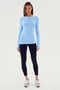 A woman stands facing forward, wearing a SPLITS59 Louise Rib Long Sleeve in Skylight, black leggings, and white sneakers. She has long, straight hair and a neutral expression. The plain white background complements her poised stance, ready for yoga or gym workouts.