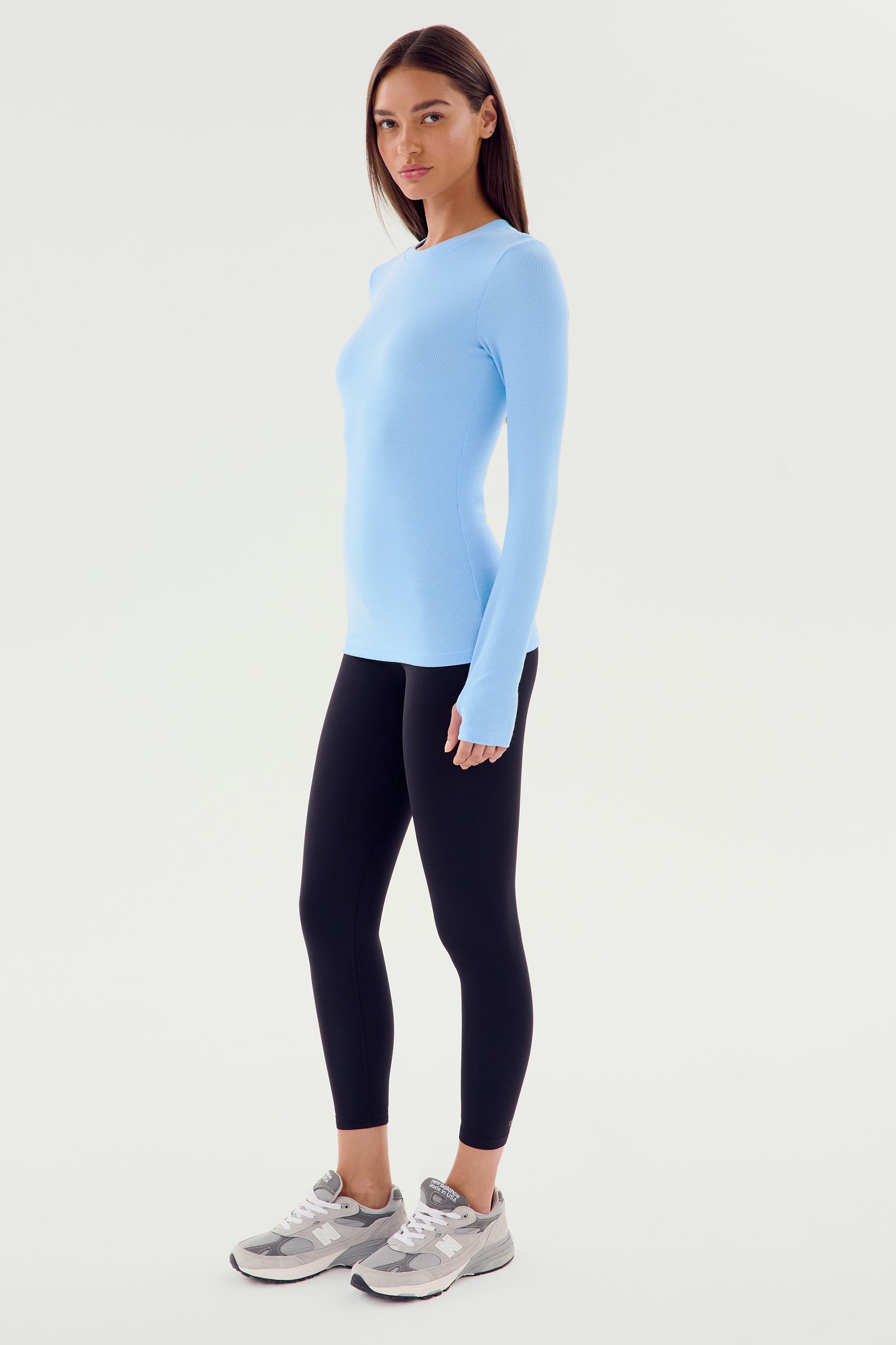 A woman in a Skylight Louise Rib Long Sleeve shirt by SPLITS59, black leggings, and grey sneakers stands in a side profile against a plain white background, looking ready for her gym workouts.