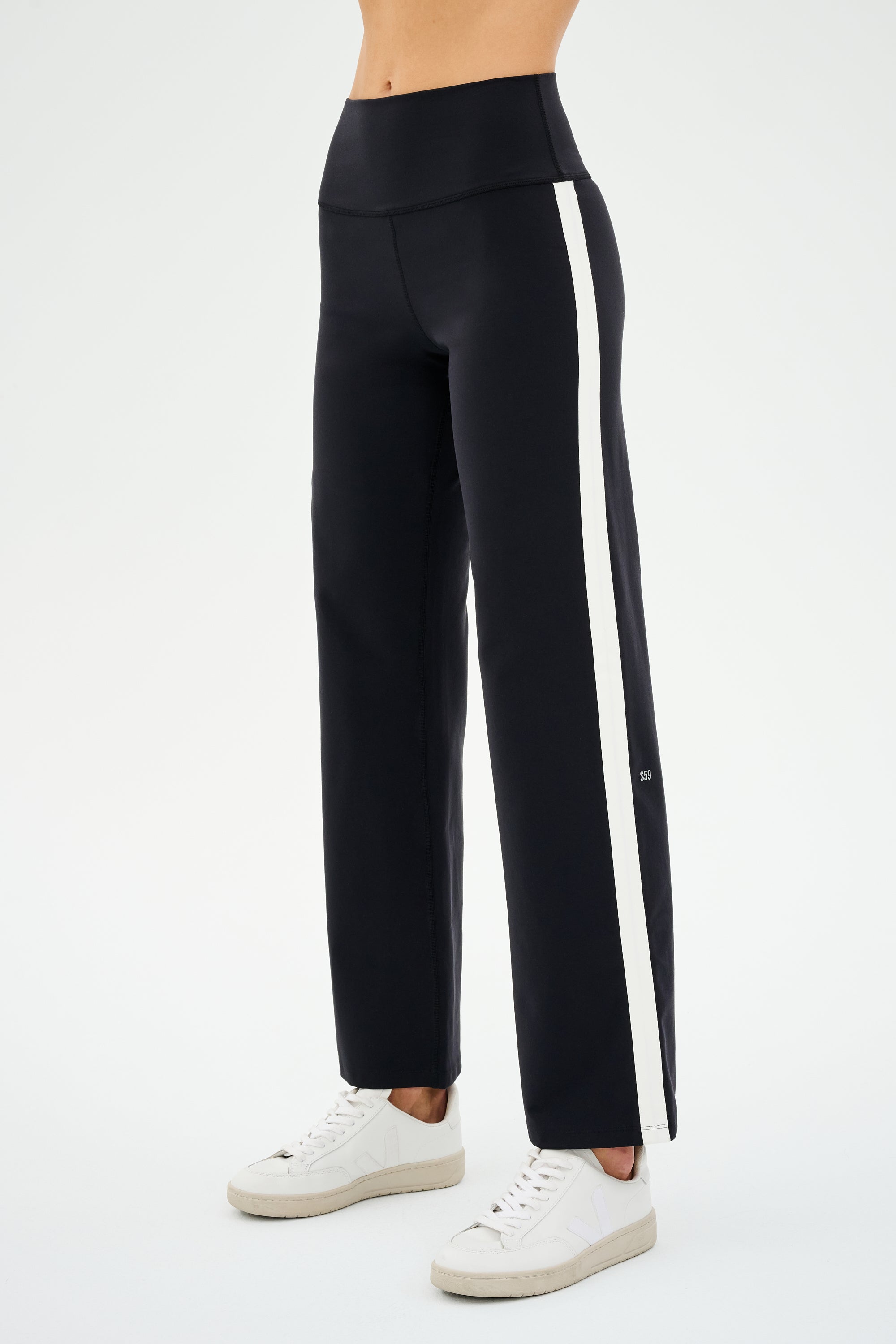 A person stands on a neutral background wearing SPLITS59's Harper Supplex Pant in Black/White with white side stripes, paired with stylish white sneakers.