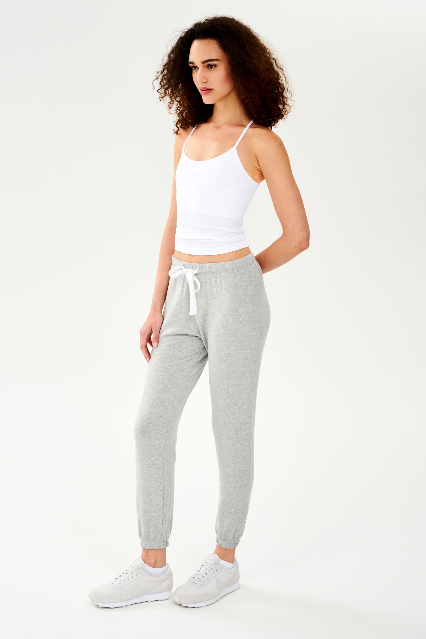 A woman wearing a SPLITS59 Loren Seamless Waist Length Tank in White with a built-in shelf bra and grey sweatpants.