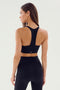 A person with long hair is seen from the back, wearing a black Sara Airweight Bra from SPLITS59 and high-waisted black leggings, perfect for hot yoga. The background is plain and light-colored.