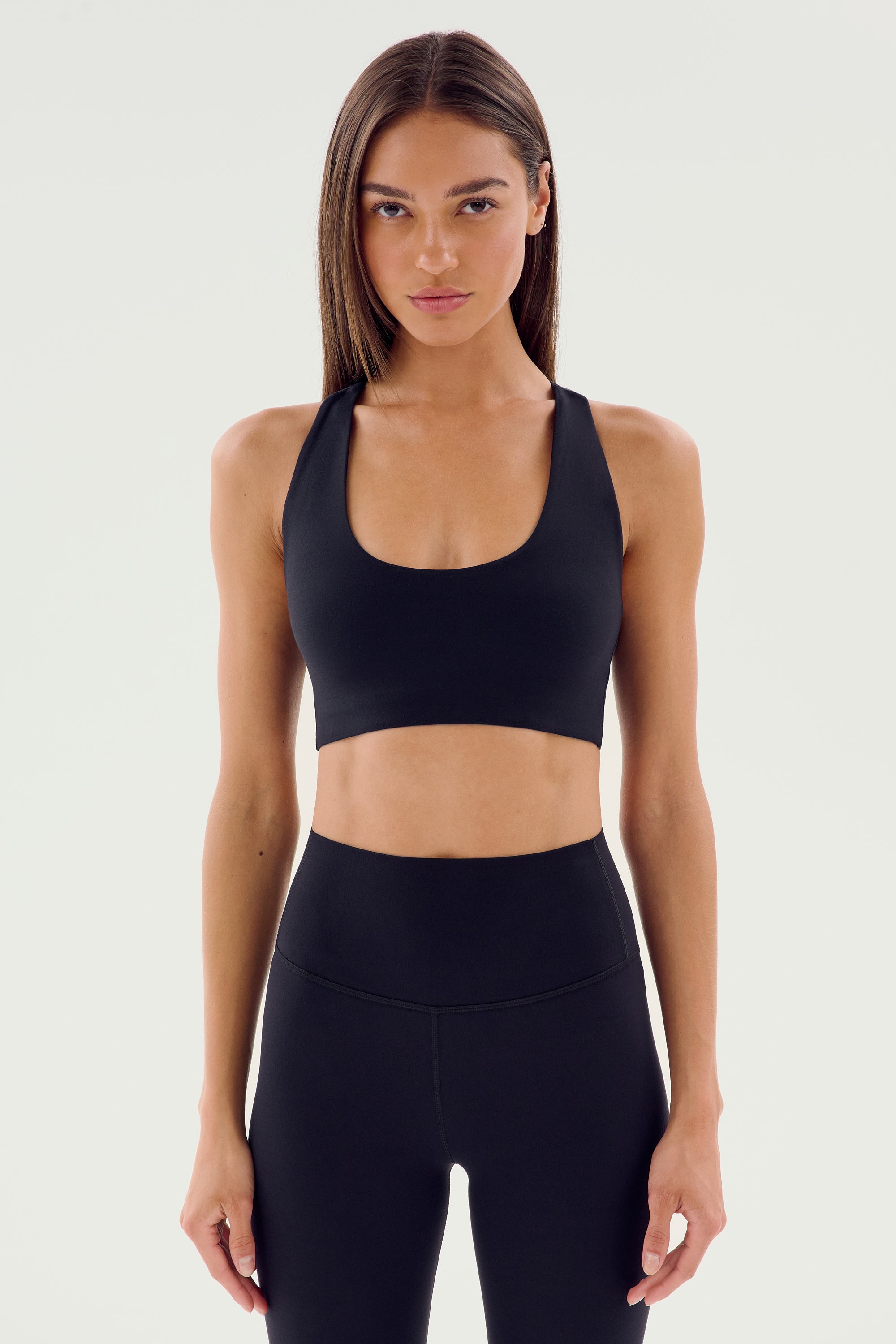 A person wearing a black SPLITS59 Sara Airweight Bra and black leggings stands facing the camera against a plain background, ready for a Pilates session or some hot yoga.
