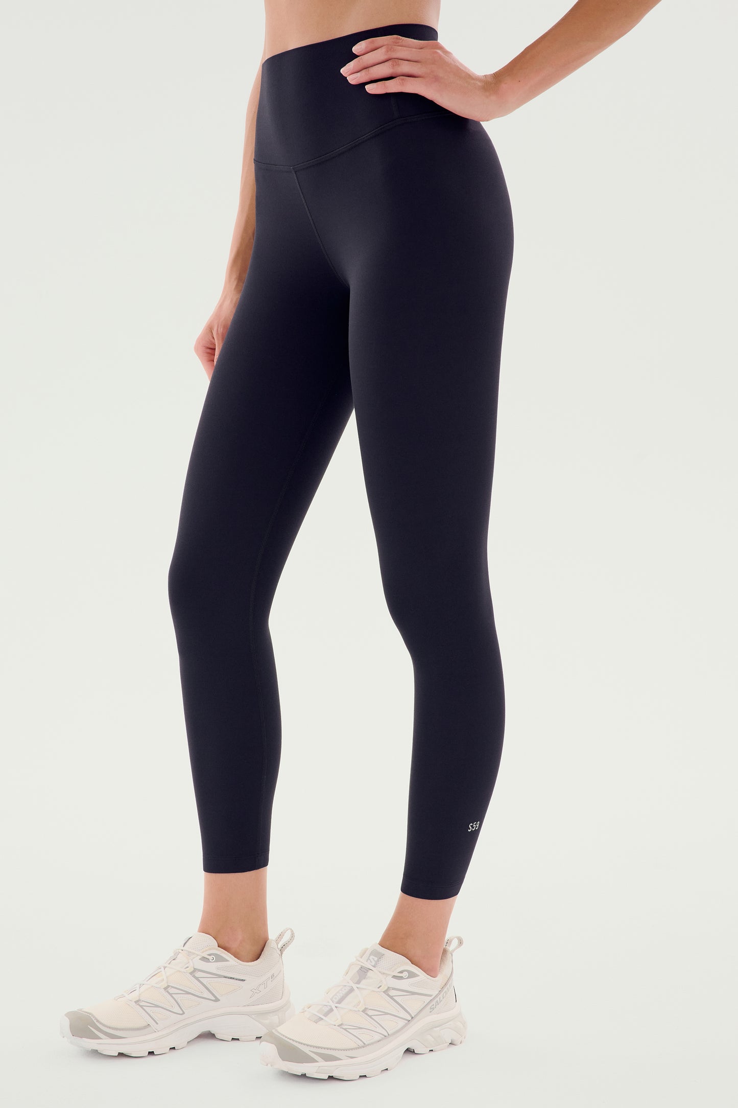 A person wears the SPLITS59 Airweight High Waist Legging in Black paired with white athletic shoes, standing against a plain light background.