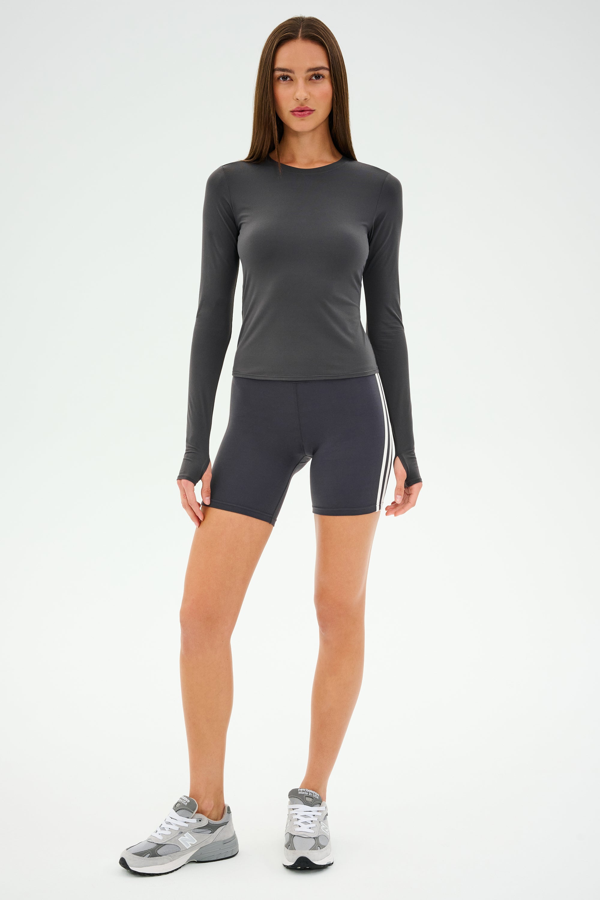 A person wearing SPLITS59's Airweight Lite LS Waist Length top in Graphite, dark side-striped shorts, and lightweight sneakers stands against a plain backdrop.