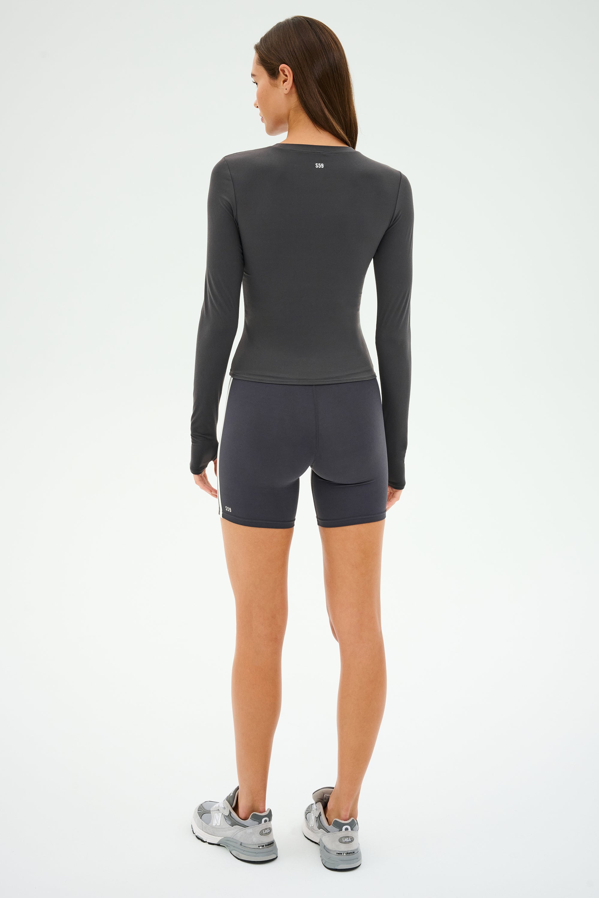A person stands with their back facing, wearing a SPLITS59 Airweight Lite LS Waist Length top in Graphite and shorts paired with sneakers on a plain background. Perfect for intense workouts!.